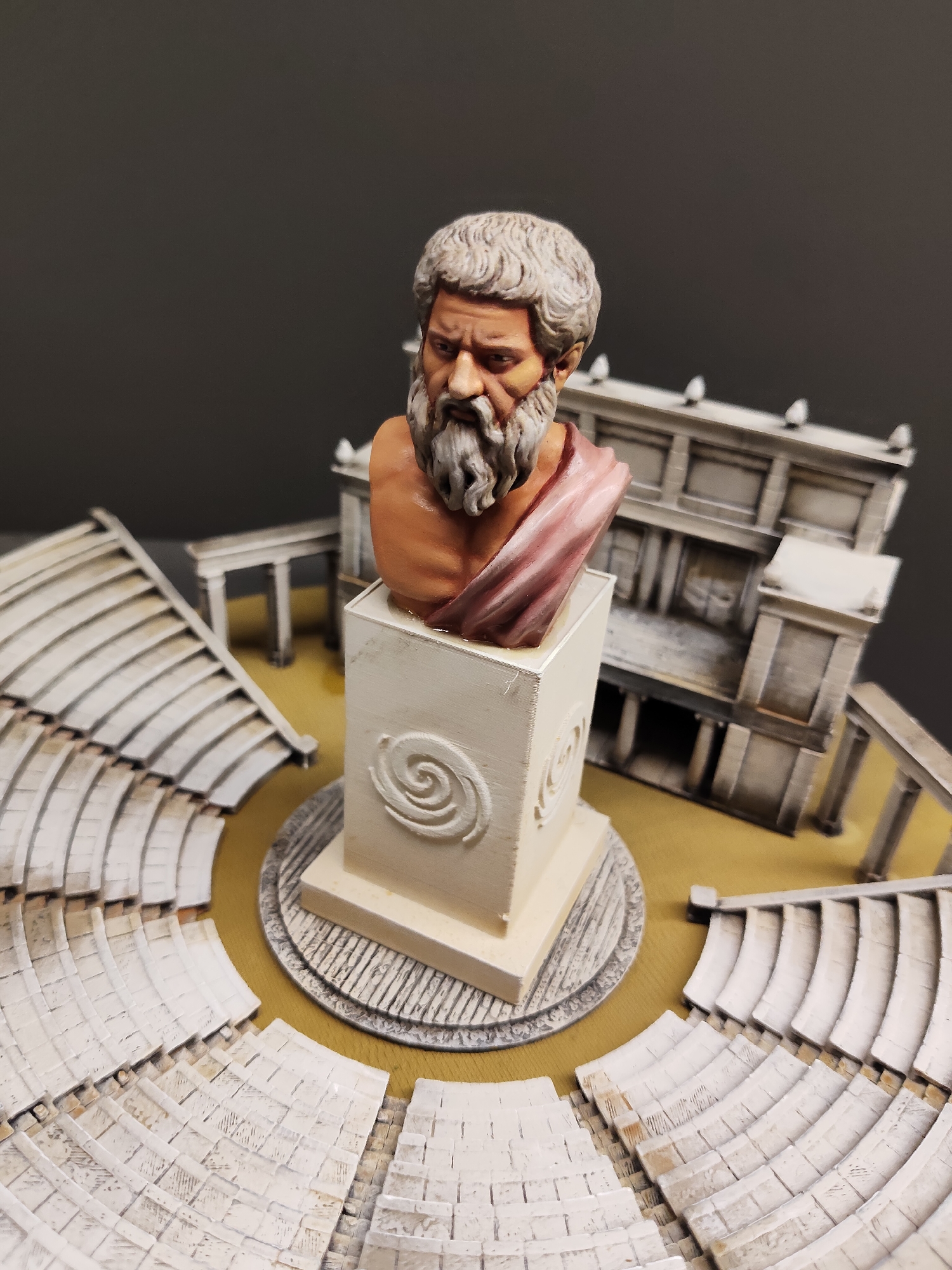 The image shows a miniature diorama of an ancient Greek theater with a bust of a bearded man, likely a philosopher or historical figure, placed on a pedestal in the center. The theater features tiered seating arranged in a semi-circular fashion and a building resembling a classical Greek facade in the background. The diorama is set against a plain dark gray background, suggesting it is a studio shot or display. The scene is lit to highlight the details of the miniature figures and architecture. There is no indication of the location being in Singapore.
