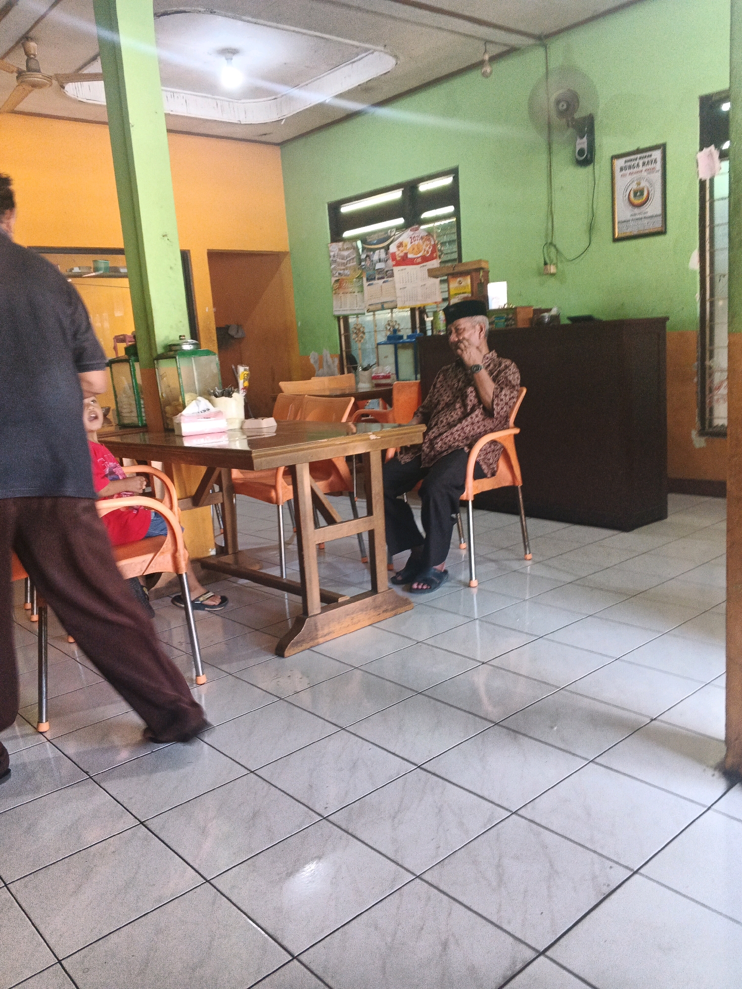 This is a medium-shot, indoor photograph of a group of people in what appears to be a simple restaurant or food stall in Ungaran, Indonesia. The walls are painted in two contrasting colors: a bright orange on the left and a light green on the right. A large, light green pillar is visible on the left side of the frame, supporting the ceiling.
In the foreground, a person wearing dark brown pants and a dark gray shirt is walking away from the camera, towards the left. Their feet are visible, as are their brown pants.
Seated at a wooden table in the center of the image is an elderly man wearing a black cap and a patterned brown and white shirt. He is gesturing with his right hand and looking to his left, smiling. He is seated on an orange plastic chair. The table is laden with a few items, including tissues in a white box, a container with utensils, and a small glass dispenser.
To the left of the elderly man, a young child, partially visible and wearing a red shirt, is seated at the same table on an orange plastic chair. The child appears to be looking towards the elderly man or the person walking away.
In the background, more orange plastic chairs and tables are visible. There is a dark brown counter or cabinet along the back wall, and on the wall, a framed certificate or sign with Indonesian text is displayed. A ceiling fan is mounted on the green wall, and a light bulb hangs from the ceiling. The floor is tiled with light-colored square tiles. The overall impression is of a casual, local eatery.