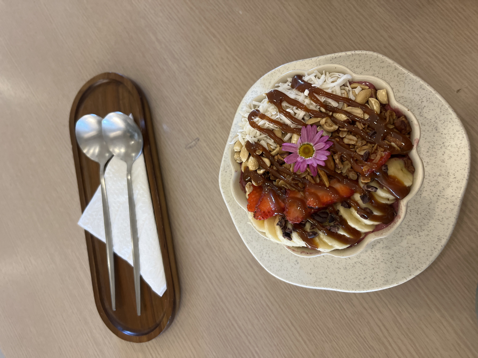 A top-down shot depicts an acai bowl, garnished with sliced strawberries, bananas, granola, nuts, shredded coconut, a drizzle of caramel, and a single purple flower. The bowl rests on a speckled, off-white plate, placed on a light-colored wooden table. To the left, a small, elongated wooden tray holds two silver spoons and a folded white napkin. This scene could be from a cafe or restaurant in Puebla, Mexico.