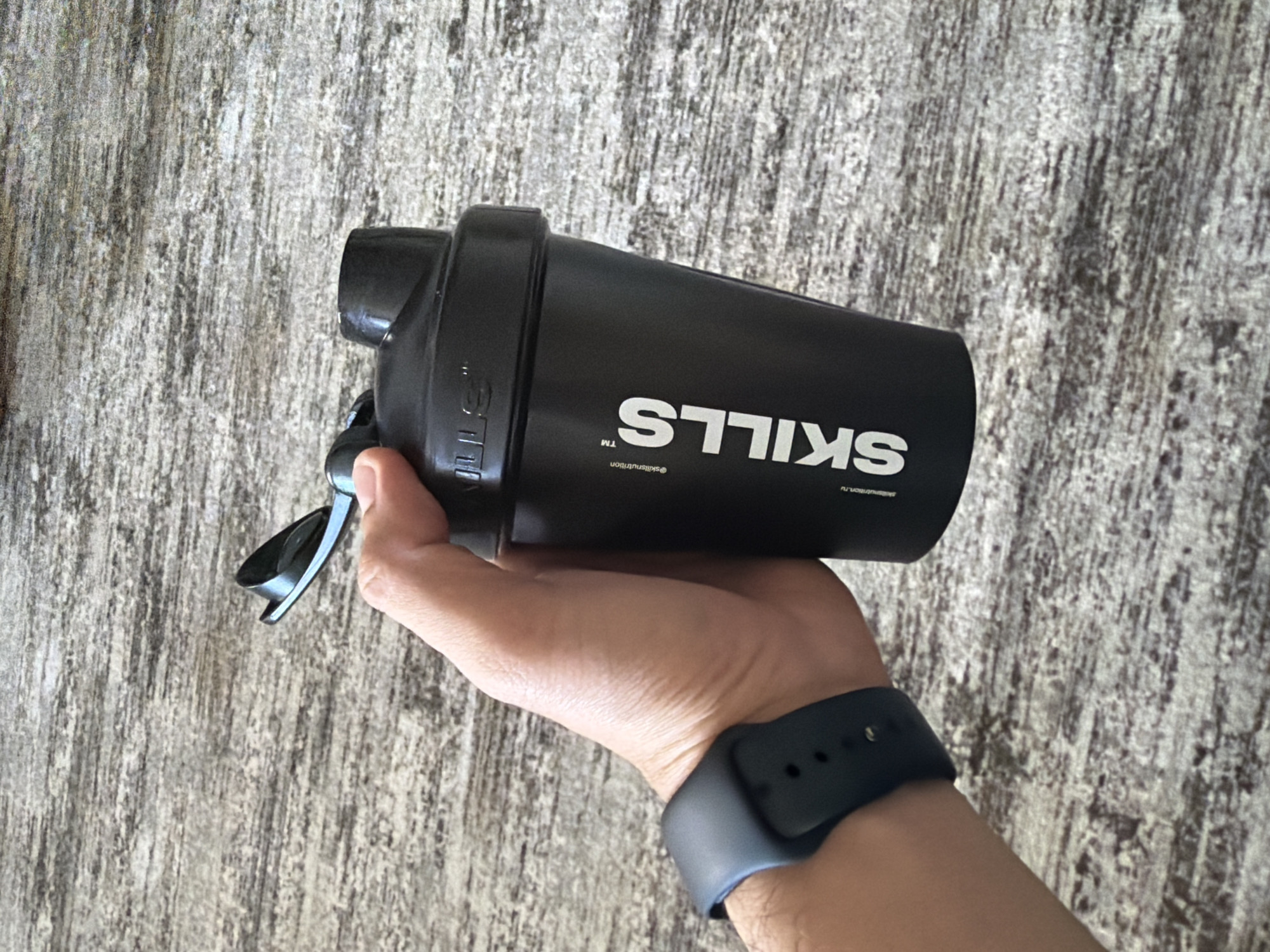 A close-up shot features a human hand holding a black shaker bottle against a light, textured background. The hand is positioned centrally, gripping the bottle firmly. A black smartwatch with a matching band is visible on the wrist.
The shaker bottle is matte black with white branding. The word "SKILLS" is prominently displayed in large, bold white capital letters, appearing upside down from the viewer's perspective. Below it, also inverted, are the smaller "™" symbol and the text "@skillsnutrition". Even smaller, below that, is partially discernible text that includes "nutrition.ru" or a similar web address, also upside down. The shaker's lid, located on the left, has a black loop or handle attached and shows some small, indistinct white markings or a logo, possibly "STK," which are also inverted.
The background is a textured surface with varying shades of light grey, white, and faint brown, featuring vertical, linear patterns, resembling distressed wood or a roughly textured wall. The lighting is bright and diffuse, creating minimal shadows. The image captures a simple action of holding the bottle. This scene takes place in Chaikovsky, Russia.