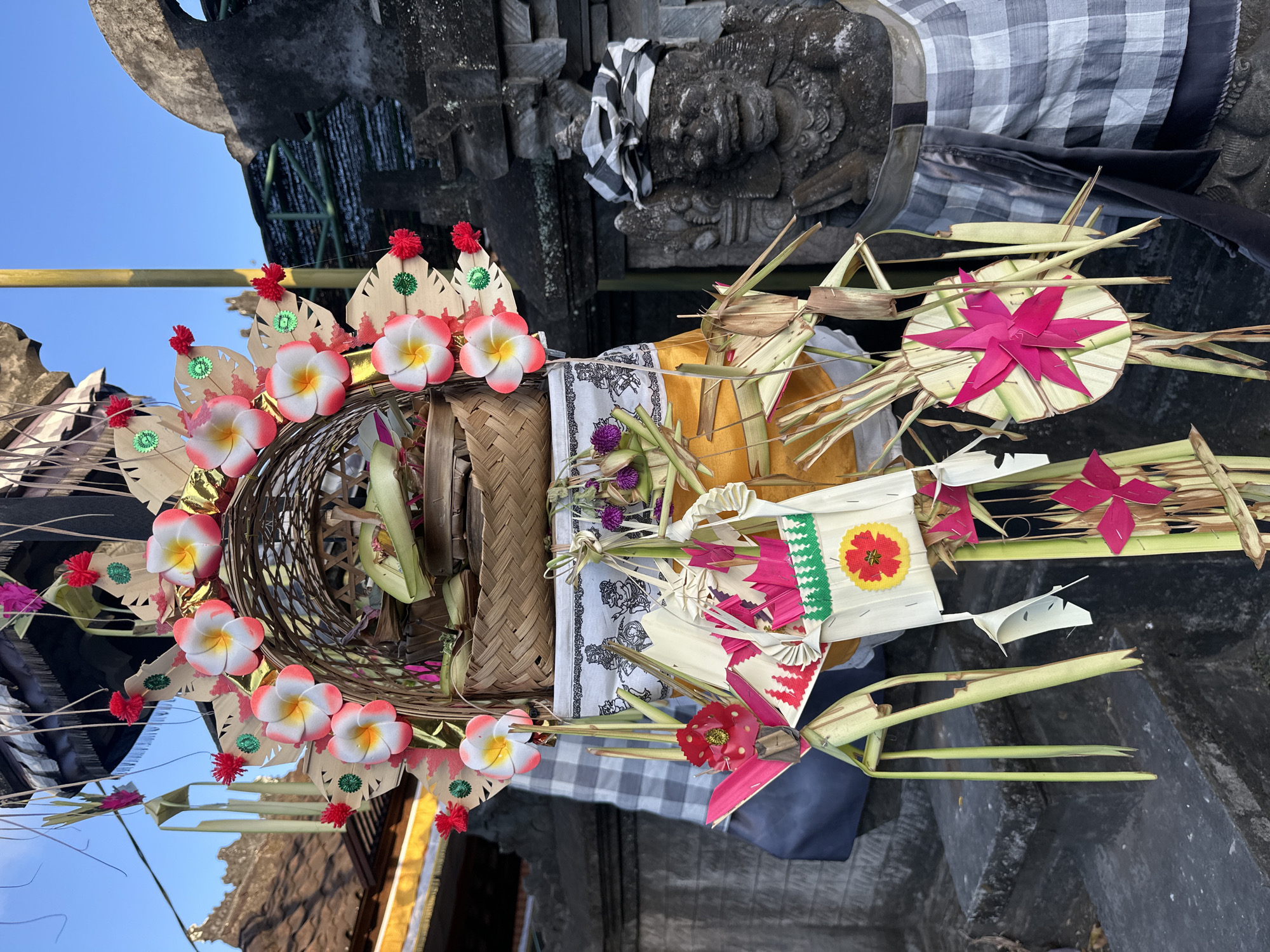 This image depicts a close-up view of a Balinese offering, likely a Canang Sari, placed in an outdoor setting. The offering is arranged in a woven basket and features a variety of decorative elements. Prominently visible are artificial plumeria flowers in white and pink hues, each with a red pom-pom and green accents. There are also folded palm leaf decorations, some with floral patterns in pink and red, and others with circular designs. Dried palm fronds are used as a base and to create a festive, natural look.
The offering is set against a backdrop of stone structures, suggesting a temple or traditional Balinese architectural site. A black and white checkered cloth, characteristic of Balinese offerings, is draped over a stone carving, possibly a deity or guardian figure. The overall scene is brightly lit, indicating it is daytime, with a clear blue sky visible in the upper left corner. The angle of the sun suggests it might be morning or late afternoon. The composition emphasizes the intricate craftsmanship and vibrant colors of the offering, highlighting its ceremonial importance. No visible text is present in the image. The location context of Kuta Selatan, Indonesia, further reinforces the cultural significance of this type of offering.