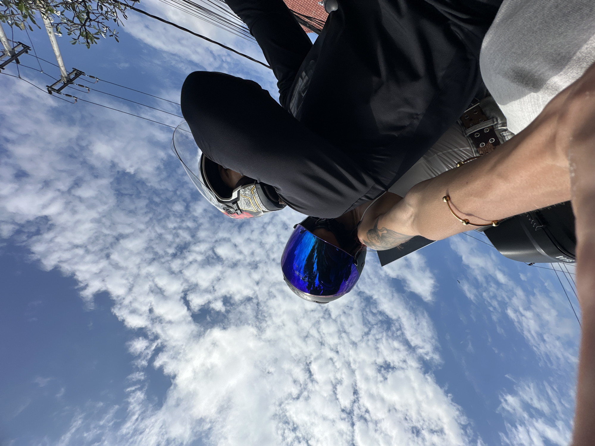 The image, taken with a tilted perspective, captures a view of the sky above Kuta Selatan, Indonesia. The sky is partially covered with white clouds against a blue backdrop, while utility poles and wires are seen along the upper left side. In the foreground, the lower half of a person is visible wearing riding gear, including a black long-sleeved shirt and white or gray pants. They are wearing a motorcycle helmet with a reflective blue visor. A portion of a tattooed arm with a gold-colored bracelet is also visible on the right side.