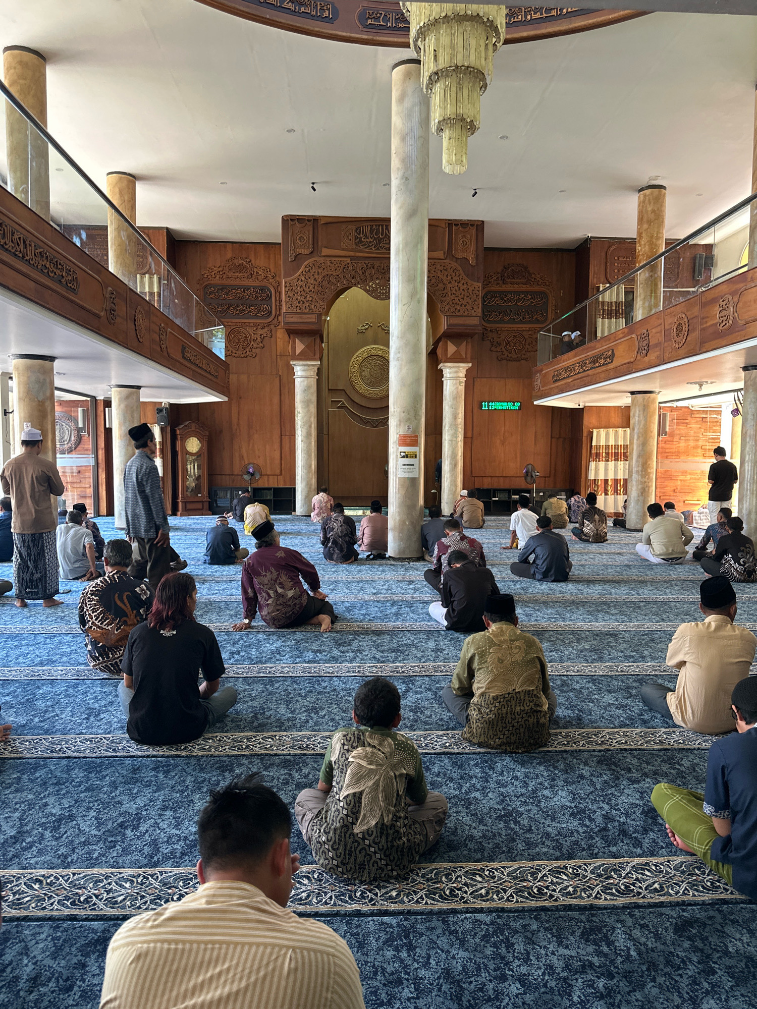 The image depicts the interior of a mosque in Malang, Indonesia, likely during prayer or a religious gathering. Approximately thirty individuals, all appearing to be adult males, are seated on a blue carpet with intricate patterns. They are facing a large wooden structure at the far end of the room, which features ornate carvings and what appears to be a mihrab. Several individuals are in prayer positions, while others are sitting in contemplation.
The mosque has a high ceiling with a decorative chandelier. Large, marbled pillars support the upper level of the mosque, which is accessible via a staircase. The walls are adorned with wooden panels and decorative calligraphy. Natural light streams in from windows, illuminating the space. A grandfather clock is visible in the background. The overall atmosphere suggests a serene and communal religious setting.