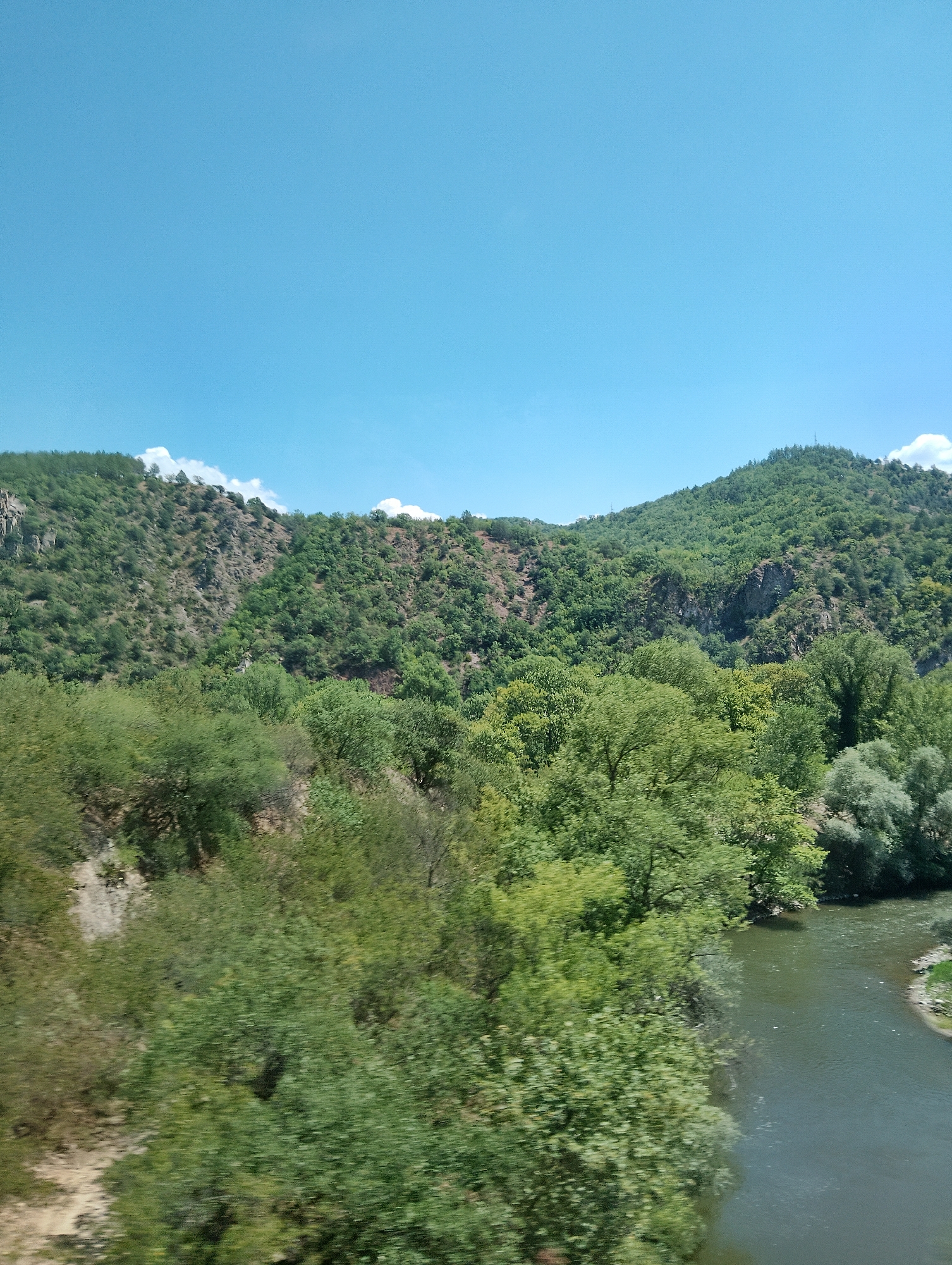 The image depicts a natural landscape with a clear blue sky above rolling green hills. The hills are covered in dense vegetation, with various shades of green indicating different types of trees and plants. A winding river flows through the lower right portion of the scene, its dark water reflecting the sky. The foreground is dominated by lush green trees, some of which appear to be in motion, suggesting the photograph was taken from a moving vehicle. There are no visible people or man-made structures, aside from what appear to be distant utility poles on the crest of the furthest hill. The overall impression is one of a tranquil, verdant environment.
