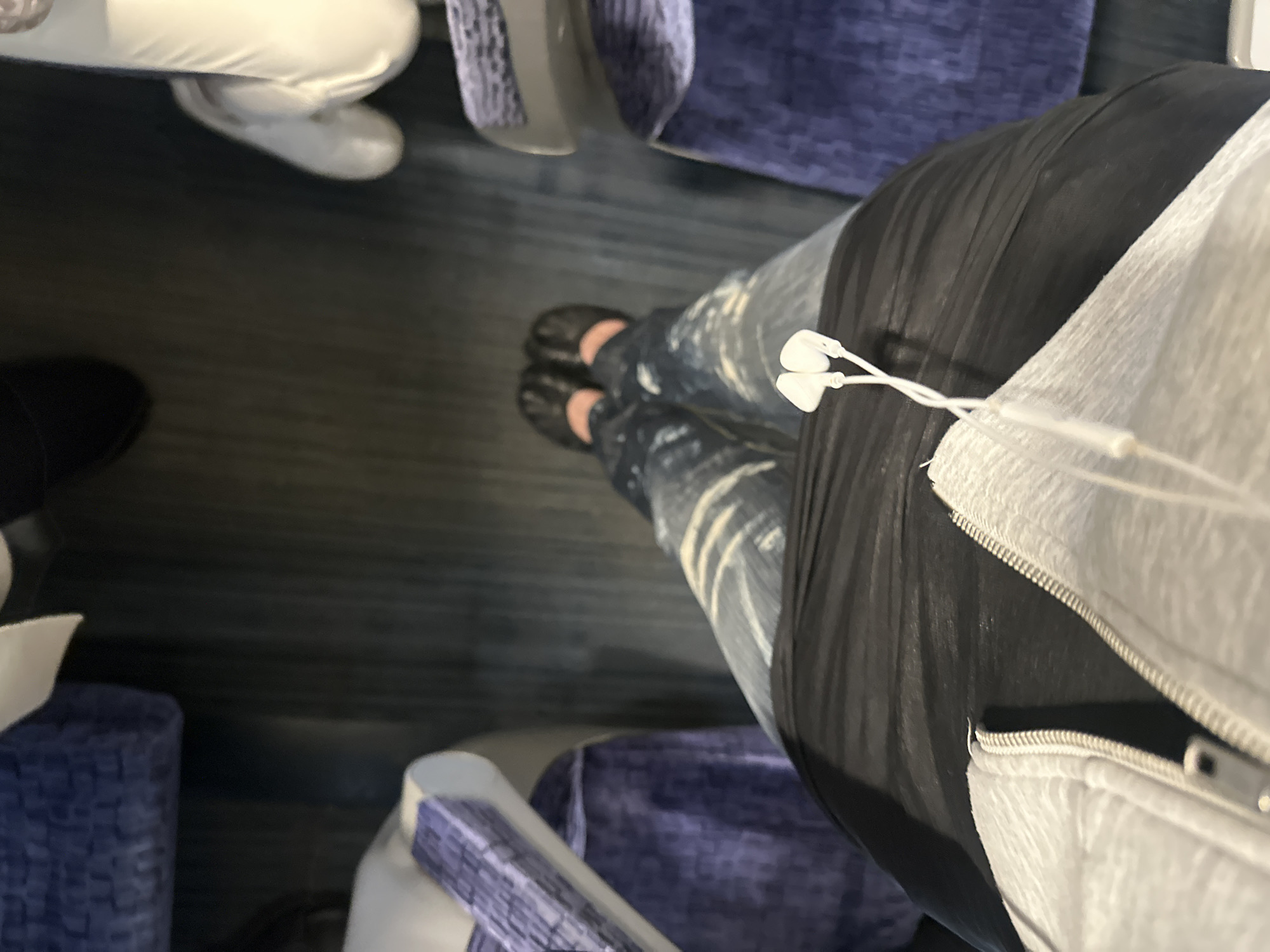 The image depicts an interior scene, likely within a public transport vehicle such as a train or bus, in London, United Kingdom, viewed from a first-person perspective looking downward. The lower half of a person is visible, wearing light-wash blue jeans, black flat shoes, and a light grey zipped hoodie over a black garment. White wired in-ear headphones hang loosely in the center foreground. The flooring is a dark blue-grey carpet with faint linear patterning. Surrounding seating structures are visible on the left and right, featuring blue/purple textured fabric upholstery with a repeating geometric design and light grey plastic or upholstered armrests/structures. In the upper right quadrant, the feet of at least one other individual, wearing dark, flat footwear and a partially visible light-colored shoe, are present on the carpeted floor.