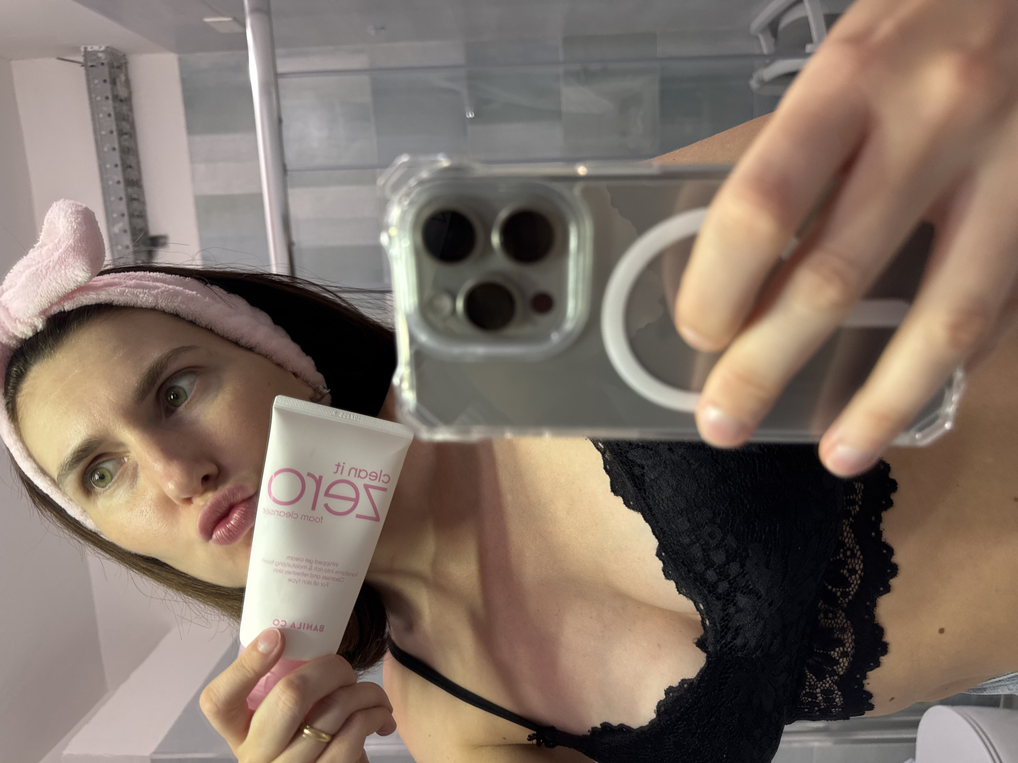 This is a selfie taken by a woman in a bathroom, likely at home. She is wearing a pink headband with rabbit ears and a black lace bra. She is holding a tube of "Clean It Zero" cleansing balm, which is partially visible with text in English and Korean. Her makeup appears to be minimal, and she is making a kissing face at the camera. The background shows a tiled wall and what appears to be a shower enclosure. The lighting suggests it is daytime. The overall mood is casual and playful.