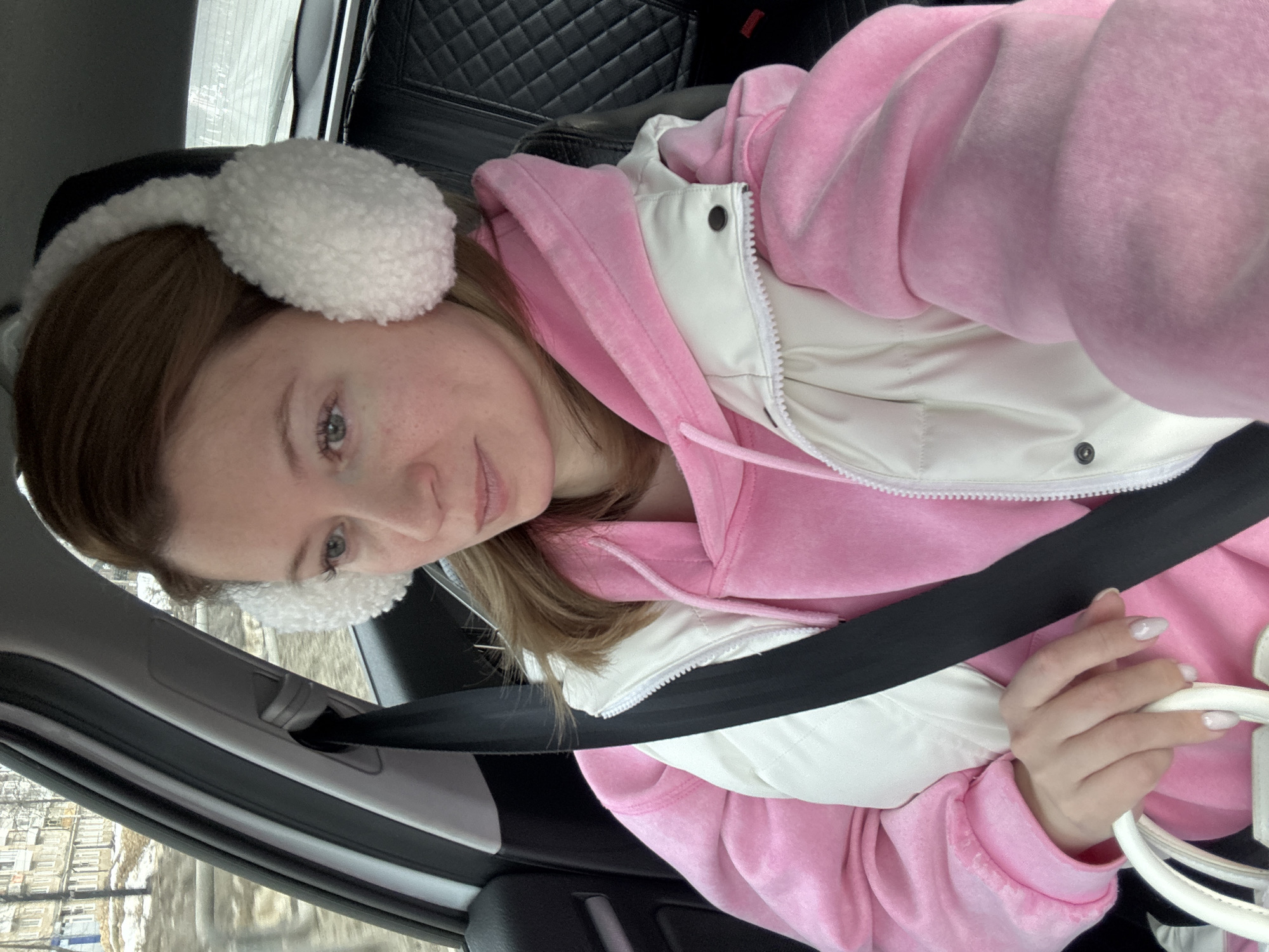 A light-skinned woman wearing white earmuffs, a pink hoodie, and a white puffer vest is seated in a car, wearing a seatbelt. She's holding a white purse. The background shows an urban landscape with buildings and snow, taken in the city of Chaikovsky, Russia.