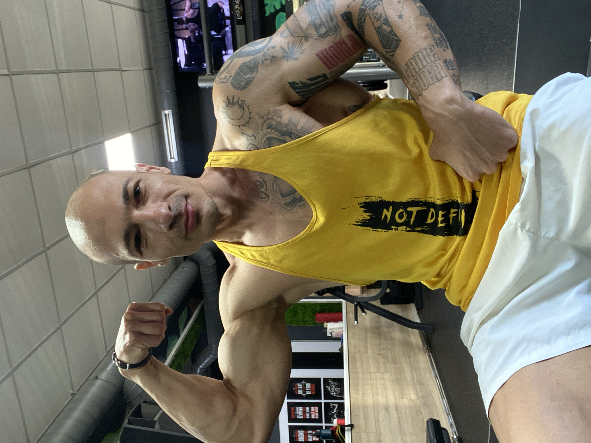 A bald man with tattoos is posing in what appears to be a gym. He is wearing a yellow tank top with the words "NOT DEFINED" printed on it and white shorts. He is flexing his left arm for the photo. The gym's equipment and interior are visible in the background. The exact location in the city of Pleven, Bulgaria cannot be determined from the image.