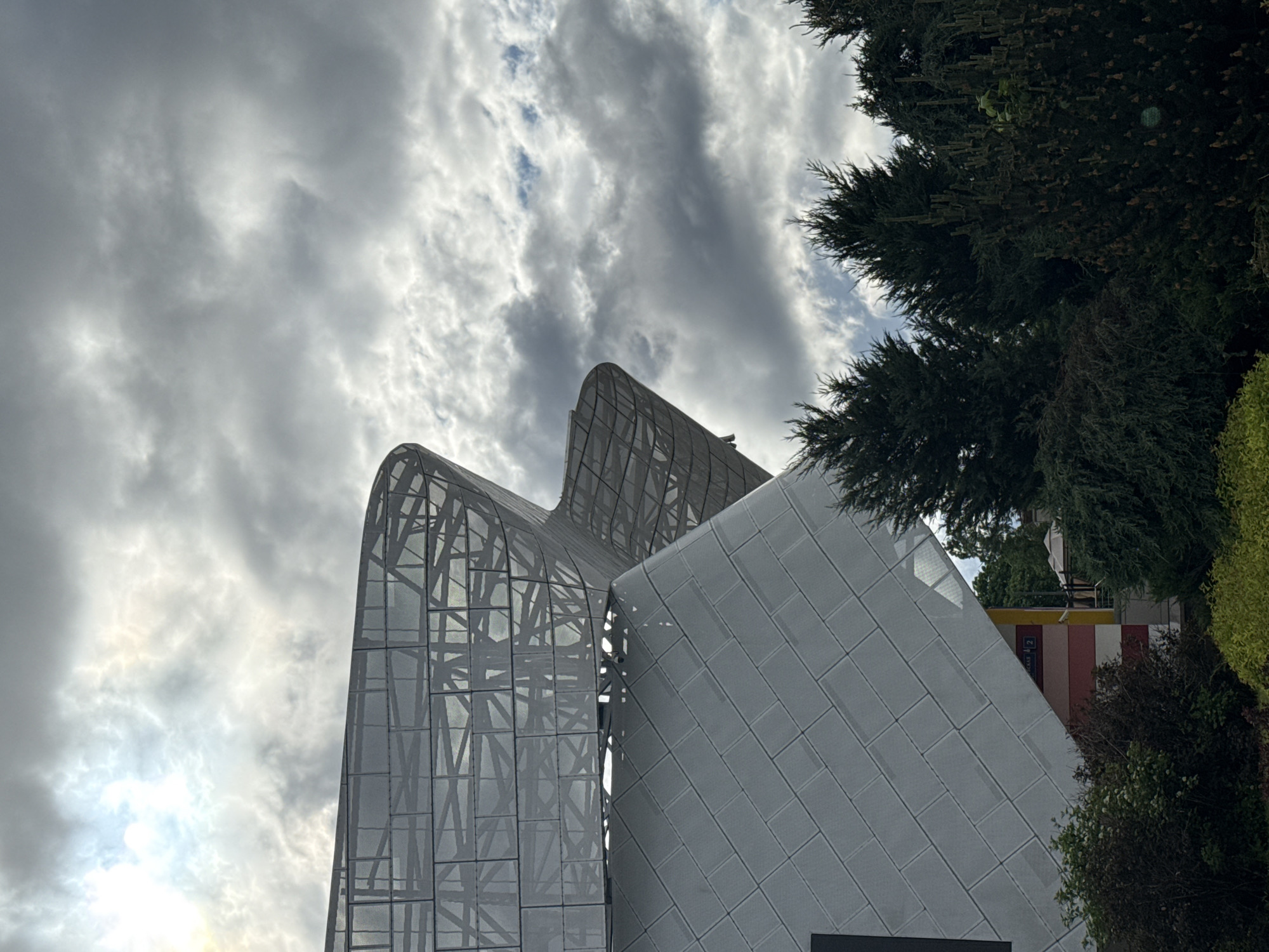The image shows a low-angle view of the exterior of the Zaryadye Park's Philharmonic Hall in Moscow, Russia. The hall is characterized by its modern architectural design, featuring curved, metal-framed glass structures. The sky is overcast with heavily clouded conditions. Green trees and shrubbery are visible on the right side of the frame, contrasting with the metallic facade of the building. In the background, a glimpse of colorful infrastructure is visible between the building and the greenery.
