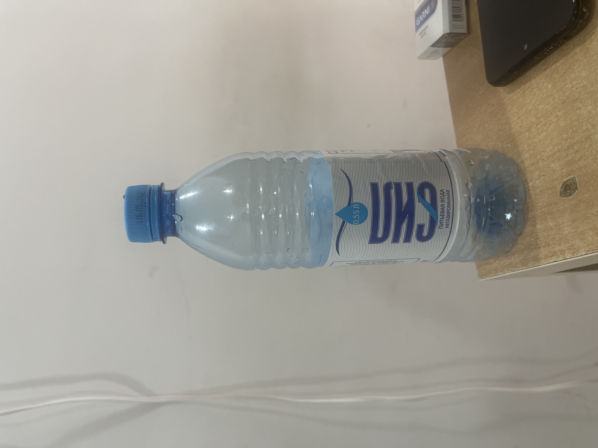 A plastic water bottle filled with clear liquid is depicted lying horizontally on a light wooden surface. The bottle has a blue screw cap and exhibits condensation or water droplets on its exterior. Its label is white and blue, featuring Cyrillic text. Visible text on the label includes "0.55 Л" (0.55 liters), "ПИТЬЕВАЯ ВОДА НЕГАЗИРОВАННАЯ" (Drinking water non-carbonated), and a large stylized word resembling "СИЛА" (SILA).
On the same wooden surface, to the upper right of the bottle, a white box with the word "GARNI" printed on it is visible, along with a partially obscured black smartphone. The background behind the objects is a plain, light-colored wall or surface. A white cable or cord runs across the bottom section of the image. The scene is static, set indoors, with no discernible activity or specific time of day indicated beyond general ambient light.
