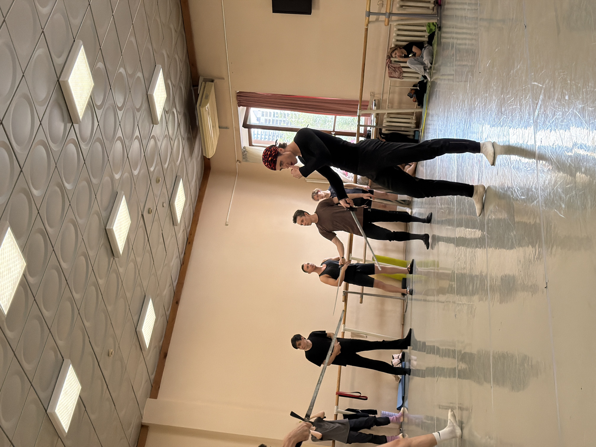 The image depicts a group of individuals in a dance studio, possibly engaged in sword fighting practice, within the city of Ufa, Russia. The studio has a polished wooden floor and light-colored walls. Several people are holding what appear to be swords or similar props, suggesting an active training session. The individuals are dressed in athletic or dance attire, including black leggings and tops, and one person wears a red bandana. A ballet barre runs along the wall, a common feature in dance studios. In the background, a window offers a glimpse of buildings outside, indicating an urban setting. Some individuals are seated or resting on the floor, while others are standing and actively participating. The lighting in the room comes from fluorescent panels on the ceiling. The overall scene suggests a practice or rehearsal space.