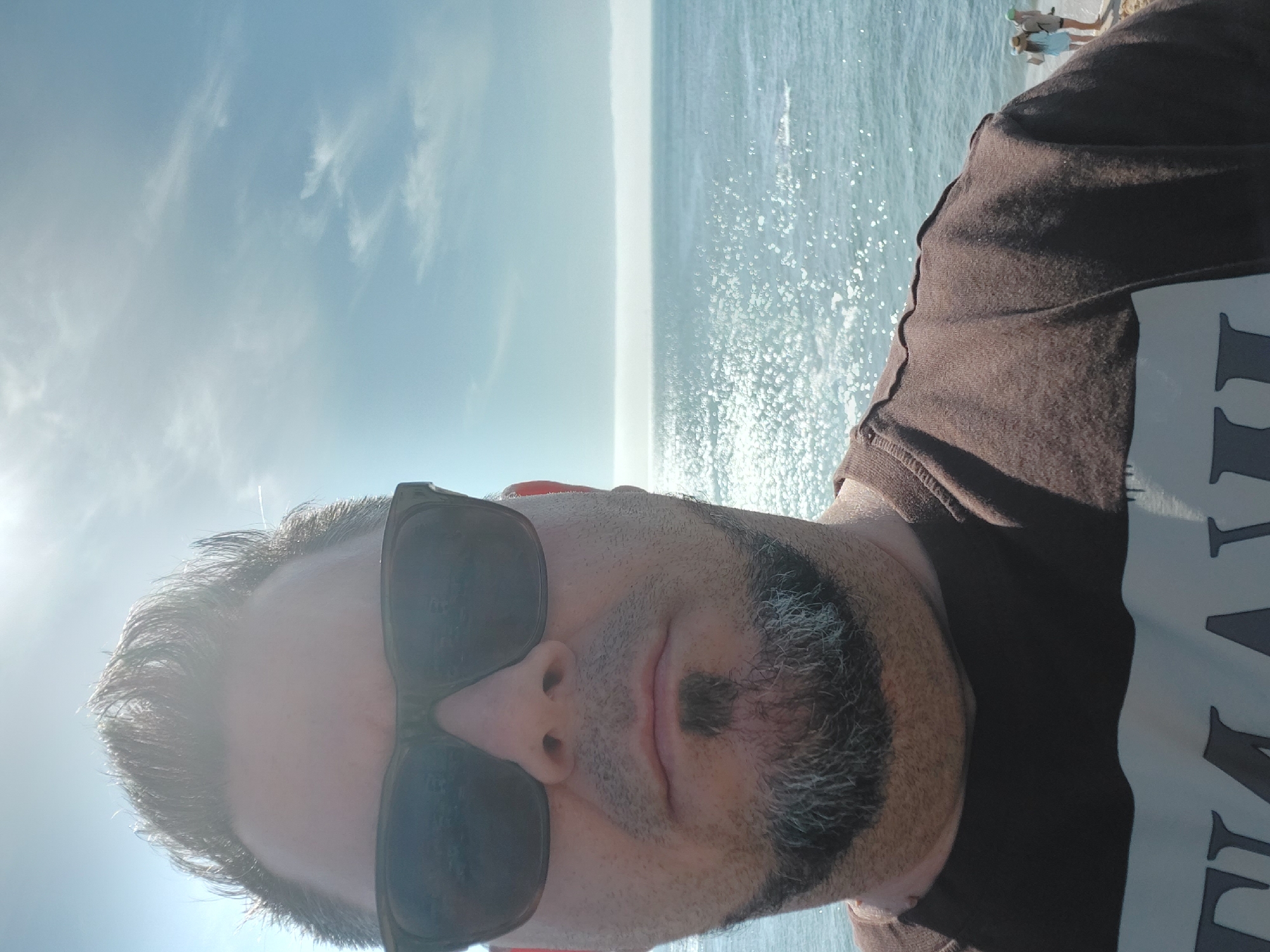 A man wearing sunglasses and a brown t-shirt is taking a selfie outdoors on a sunny day. The background shows the sea with sunlight reflecting on the water and a clear sky with scattered clouds. In the distance, two people can be seen standing near the edge of what appears to be a pier or a similar structure. Given the location specified, this scene is likely from Figueira da Foz, Portugal, a coastal city.