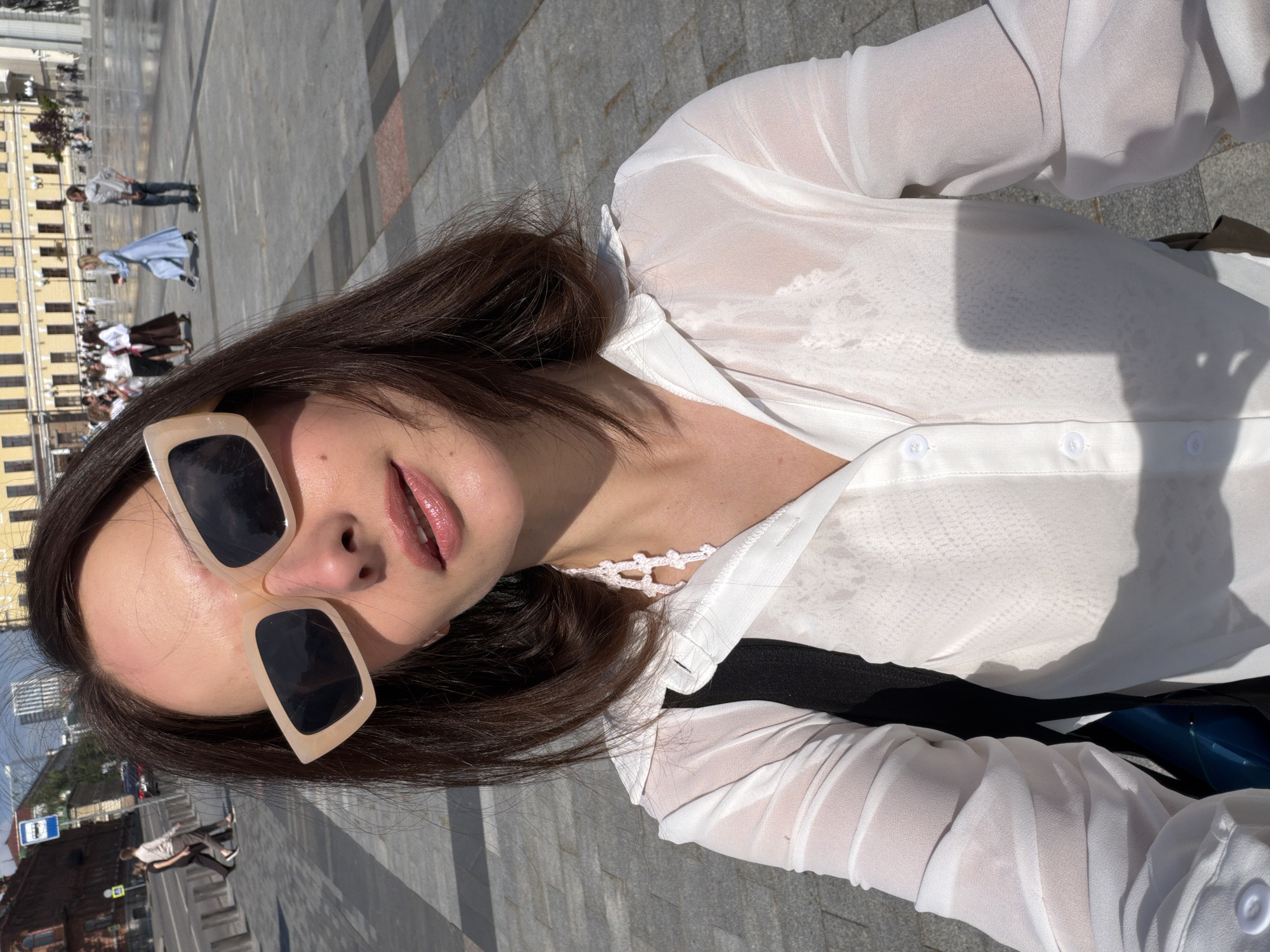 A woman with brown hair and sunglasses takes a selfie in a city square. She is wearing a sheer white blouse and a black bag is visible over her shoulder. In the background, there are buildings, people walking, and a fountain. The scene appears to be in a public urban space during the daytime.