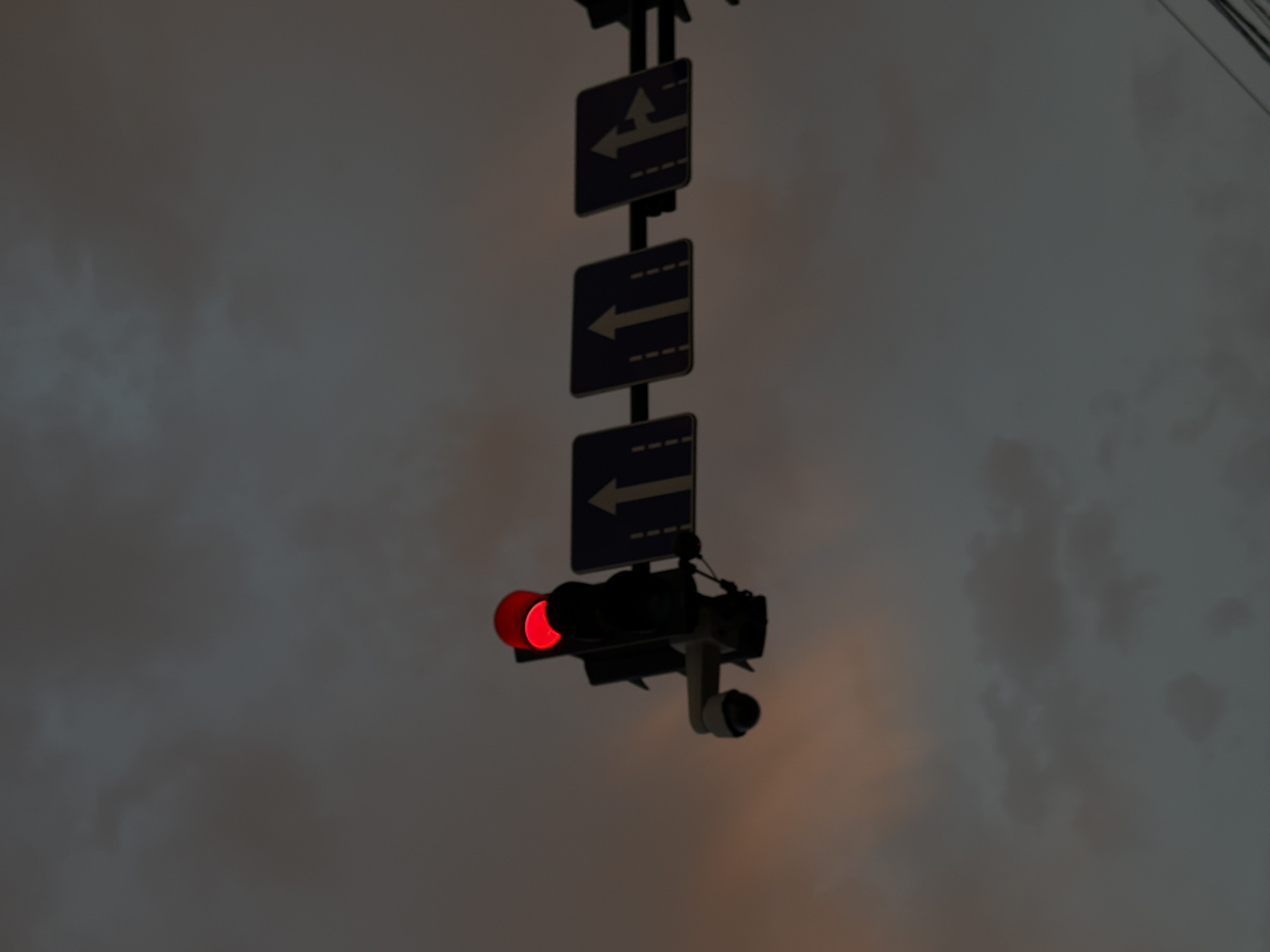 A traffic light displaying a red signal is mounted on a pole above a street in Tbilisi, Georgia. Attached to the pole below the traffic light is a surveillance camera. To the right of the traffic light, a series of traffic signs are visible. These signs indicate lane directions: one sign shows a single upward arrow, another shows two upward arrows, and a third shows an arrow pointing upward and another branching off to the right. The sky above is overcast with dark clouds, tinged with a faint orange hue from a setting or rising sun. The overall scene is dimly lit, suggesting either dusk or dawn.
