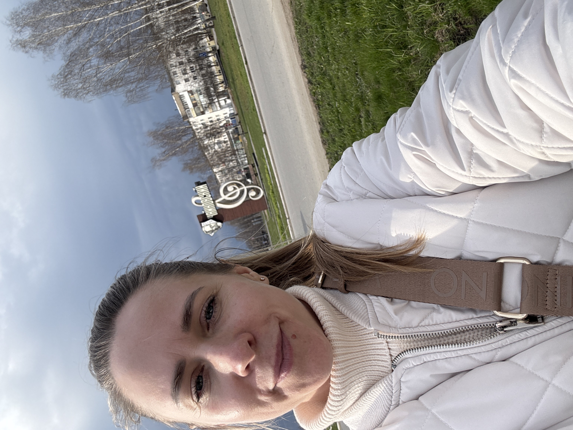 A woman takes a selfie outdoors on a clear day in what appears to be Chaikovsky, Russia. She is positioned in the foreground, smiling faintly and looking directly at the camera. Her hair is light brown, pulled back, and she has visible eyelashes and eyebrows. She is wearing a cream-colored ribbed turtleneck sweater and a light beige or off-white quilted jacket. A brown strap with the word "MONINI" visible in raised letters crosses her chest.
The background shows an urban or park-like setting. To her left and behind her, the clear blue sky dominates the upper portion of the background. Below the sky, there are numerous bare-branched trees, possibly birches, suggesting early spring or late autumn. Behind the trees, several multi-story residential buildings are visible. In the middle ground, a paved road or pathway runs parallel to a strip of vibrant green grass.
A notable object in the mid-ground is a large, decorative sculpture featuring a treble clef musical symbol. To the left of the treble clef, Cyrillic text is visible on the sculpture, including the word "ЧАЙКОВСКИЙ" (Chaikovsky), which corresponds to the provided location context. The bright daylight and clear sky indicate a sunny day. The overall impression is one of contentment in an outdoor urban setting with a strong cultural reference.
