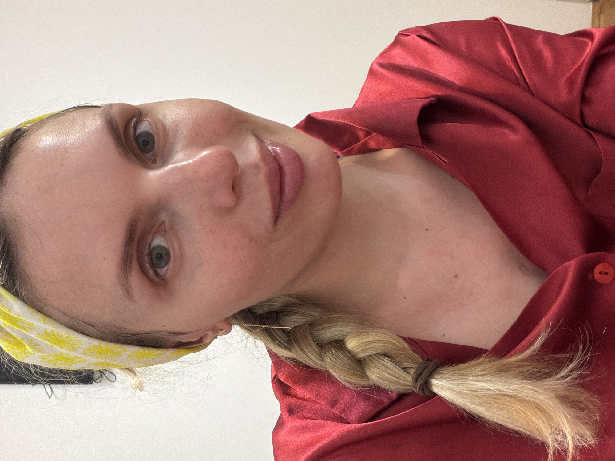 A fair-skinned woman with light blonde hair braided and tied with a brown hair tie, is wearing a yellow headband with white floral prints. She is wearing a red satin garment, possibly a robe or shirt, that is slightly open at the chest. She has light eyes and pink lips. The background is a light beige or white color. The location, Budva, Montenegro, is not discernible from the given visual information.