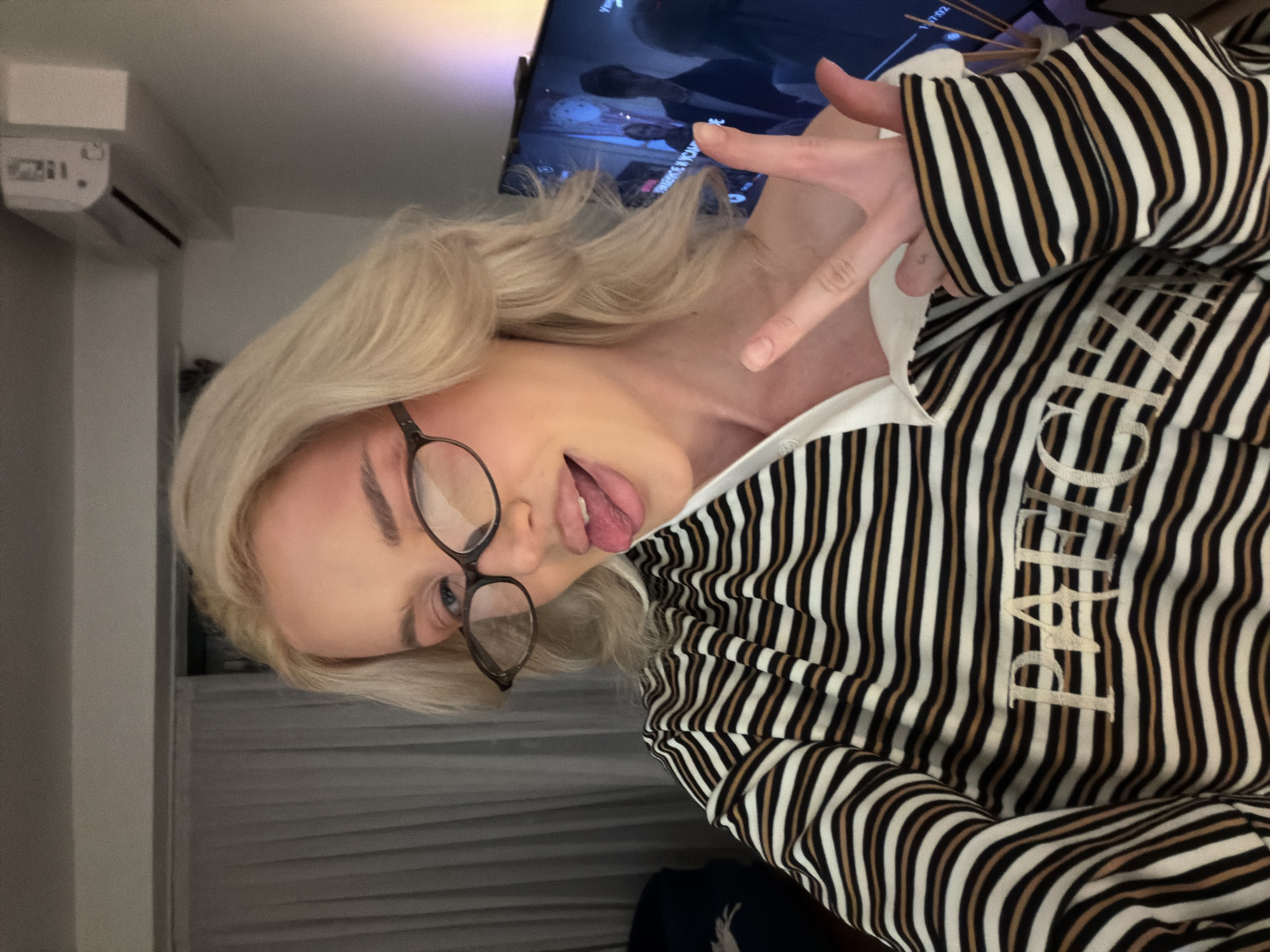 A young woman with blonde hair, wearing glasses and a striped shirt, is sticking her tongue out and holding up two fingers in a peace sign. She appears to be indoors, possibly in a residential setting. In the background, a television screen is visible, showing figures. The city and specific location within Belgrade, Serbia cannot be determined from the image.