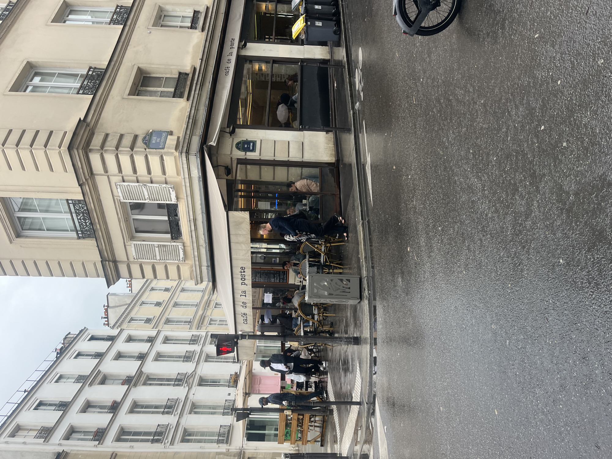 The image captures a street scene in Paris, France, likely on a rainy day, as evidenced by the wet pavement. The "Cafe de la Poste" occupies the corner of a building, with its awning and outdoor seating. Patrons are seated at the cafe, while pedestrians walk by the crosswalk. The architecture features typical Parisian buildings with multiple windows and ornate details. A street sign is visible on the corner building. A bicycle is parked on the sidewalk and a traffic light is above the crosswalk.