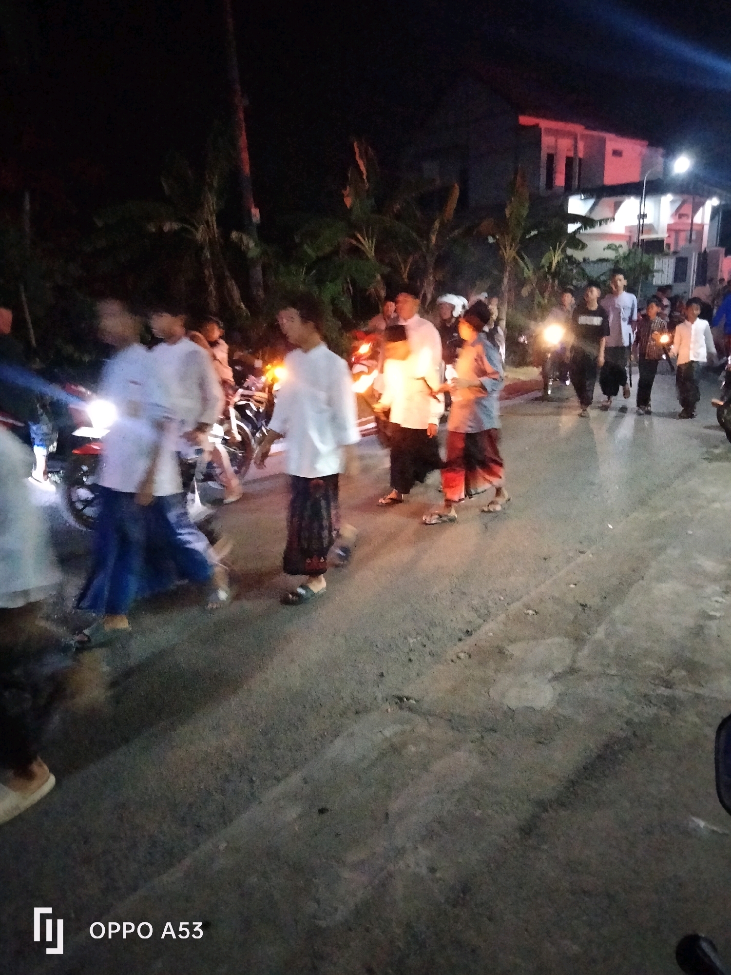 The image captures a nighttime scene on a street in Bendorejo, Indonesia, with several people gathered. The environment appears to be a residential or semi-urban area, indicated by the presence of a building with illuminated windows in the background. The lighting suggests it is after dusk, with artificial lights from buildings and vehicles illuminating the scene.
In the foreground and midground, a group of individuals, predominantly young men and boys, are visible. Many are wearing traditional Islamic attire, such as white shirts and sarongs, or more casual clothing. Some are standing and observing, while others appear to be in motion, possibly walking or interacting. There are also motorcycles present, with their headlights on, contributing to the illumination and suggesting a mode of transport or gathering point.
The people's faces are largely indistinct due to the low light conditions and the quality of the image, but their postures suggest a communal gathering. The ground is paved, appearing to be a road or pathway. The overall atmosphere seems to be that of a social event or a casual evening assembly in the community.