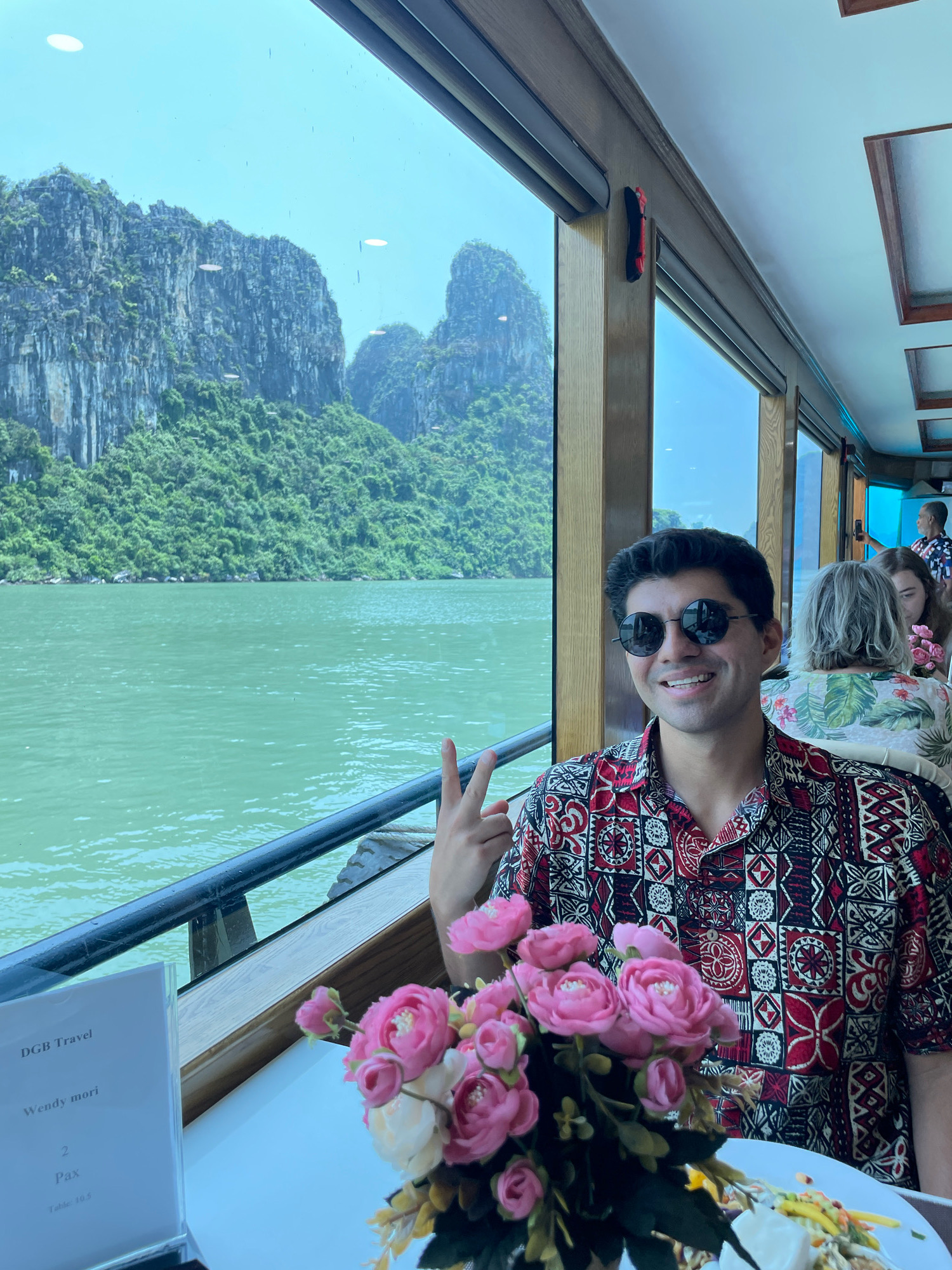 A man in a patterned shirt and sunglasses smiles at the camera, holding up two fingers in a peace sign. He is seated at a table on a boat with large windows offering a view of Halong Bay in Quang Ninh City, Vietnam. The bay is characterized by karst rock formations covered in lush green vegetation. The water is a vibrant turquoise. Inside the boat, other passengers are visible, as well as a vase of pink and white flowers on the table. A menu holder with "DGB Travel" printed on it is also on the table.