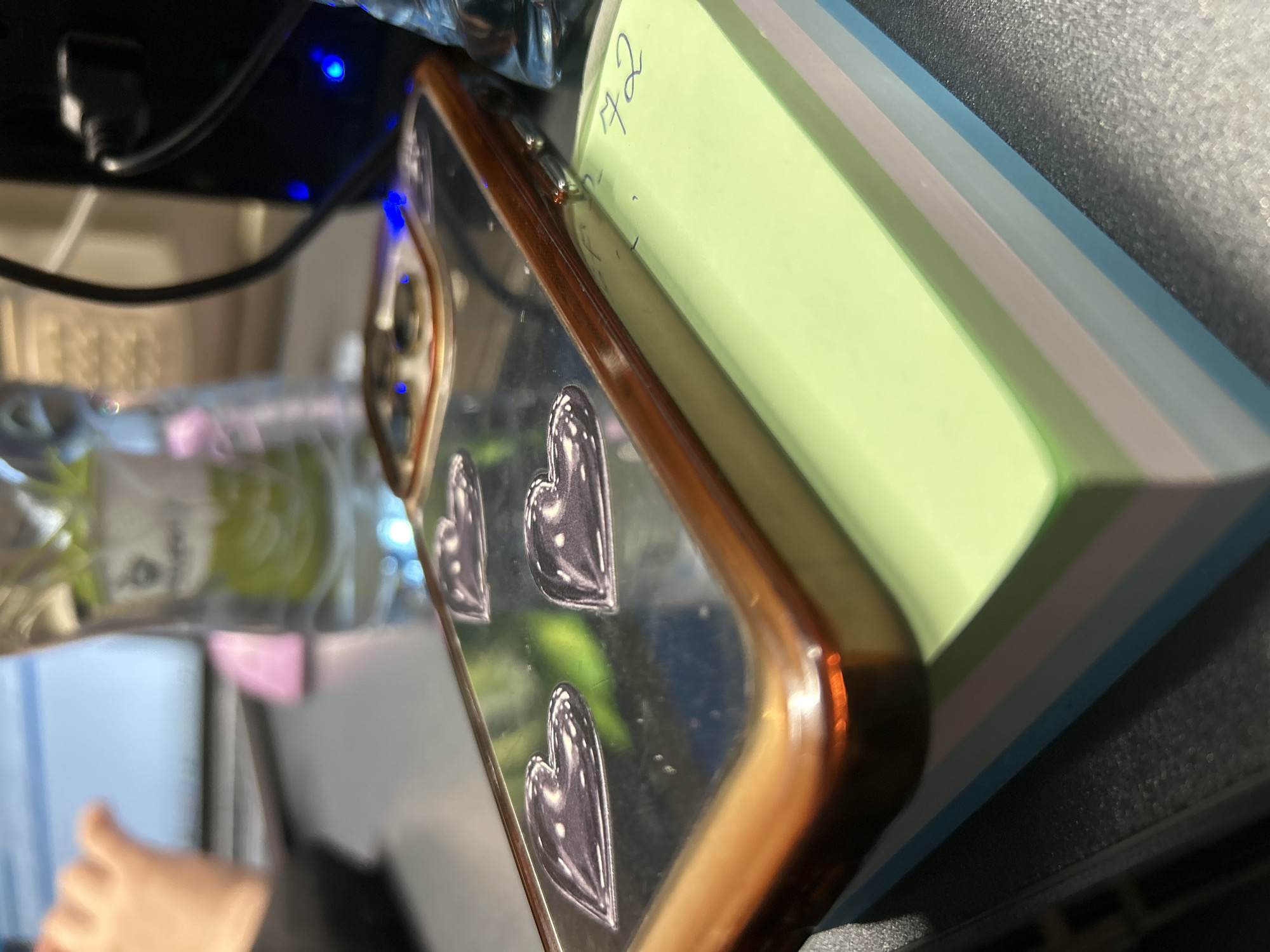 The image shows a close-up of a phone case with heart designs on a stack of colorful sticky notes. The phone case is on top of the sticky notes, which are arranged in a stack. There are some reflections on the phone case. In the background, there are some blurry objects including a bottle and what appears to be a computer or monitor. The overall scene is likely indoors, on a desk or table. There is no indication of the city of Tbilisi, Georgia, or any specific urban location.