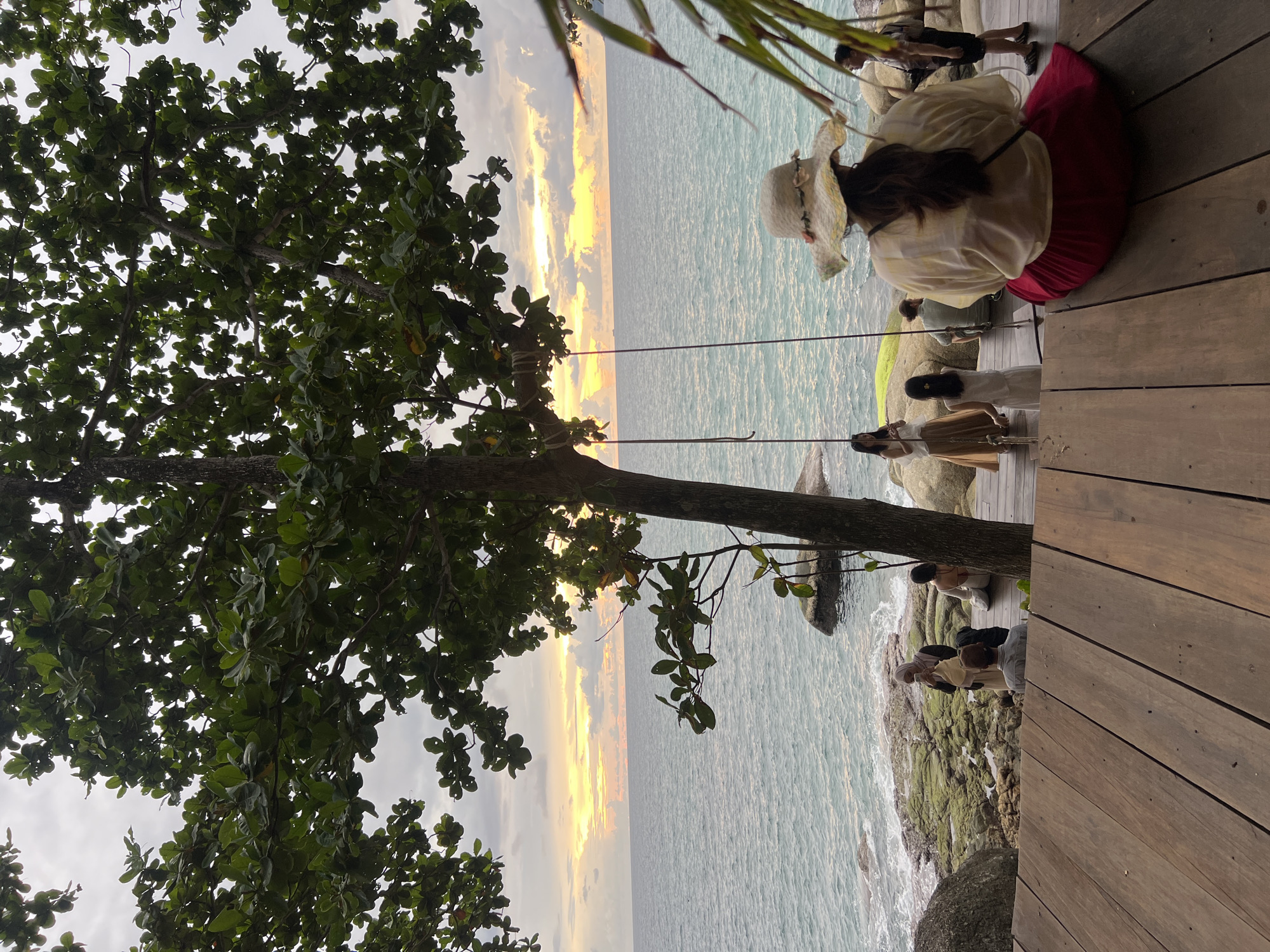 An outdoor scene at sunset on a rocky coastline in Karon, Thailand features a group of people enjoying the view. In the foreground, a wooden platform is visible with several individuals seated and standing. A woman wearing a wide-brimmed hat and a red skirt is the closest subject, facing away from the camera. To her left, other people are gathered on a wooden walkway near the water's edge.
The middle ground shows the rocky shoreline with the ocean stretching to the horizon. The water is a gentle blue-green, and waves lap against the rocks. Several large boulders are present on the shore, some covered in green moss. A tree with lush green leaves partially obscures the view of the sky and the sea.
The background displays a dramatic sunset with the sky filled with warm hues of orange, yellow, and pink, tinged with gray clouds. The sun is setting over the ocean, casting a glow on the water and the clouds. The overall atmosphere is serene and picturesque, capturing a moment of relaxation by the sea.