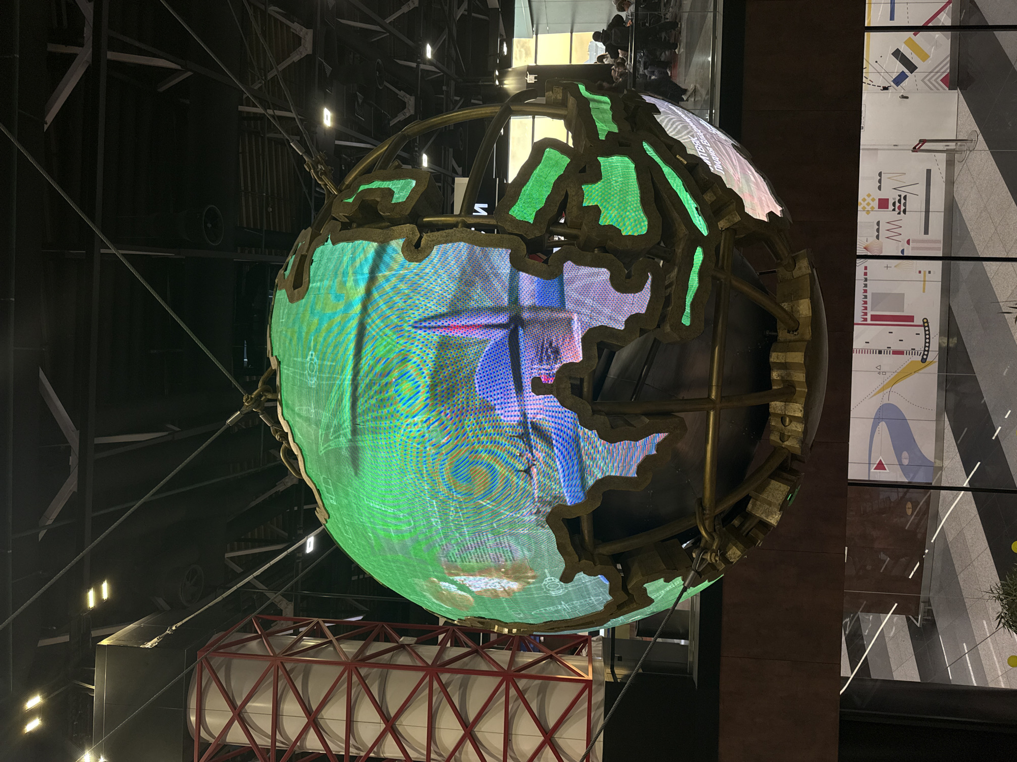 A large, illuminated spherical display, resembling a globe, dominates the center of the image. Its structure features a dark, solid core encased in a golden or bronze skeletal frame that outlines geographical landmasses. The sphere's surface is an LED screen, dynamically displaying vibrant, abstract patterns in greens, blues, purples, and yellows, some appearing as swirling graphics or technical schematics. Cables and wires are visible, suspended around the globe, connecting it to the surrounding environment.
The setting appears to be a modern, expansive indoor space, possibly an atrium or exhibition hall, characterized by industrial architecture. In the background, dark metal beams, girders, and structures are visible, with scattered rectangular light fixtures providing illumination. To the right of the globe, a tall, dark pillar stands next to a large glass wall or window. Through this glass, bright white panels adorned with colorful, abstract geometric designs (including reds, blacks, yellows, blues, and greys) are visible. Blurry figures of people can be faintly seen in the upper right background, suggesting a crowd or visitors on a higher level. Below the globe on the left, a structure with a red lattice-like frame and white segmented panels is partially visible.
The scene suggests an ongoing exhibit or public art installation, with the dynamic visuals on the globe being the primary activity. The lighting indicates an indoor setting, possibly at night or in a deliberately dim environment to enhance the glow of the display. No discernible text is clearly readable on the globe or the surrounding panels, although some patterns might resemble letters or technical drawings. The overall impression is one of modern technology and artistic display in a public urban setting, consistent with its location context in Khimki, Russia.