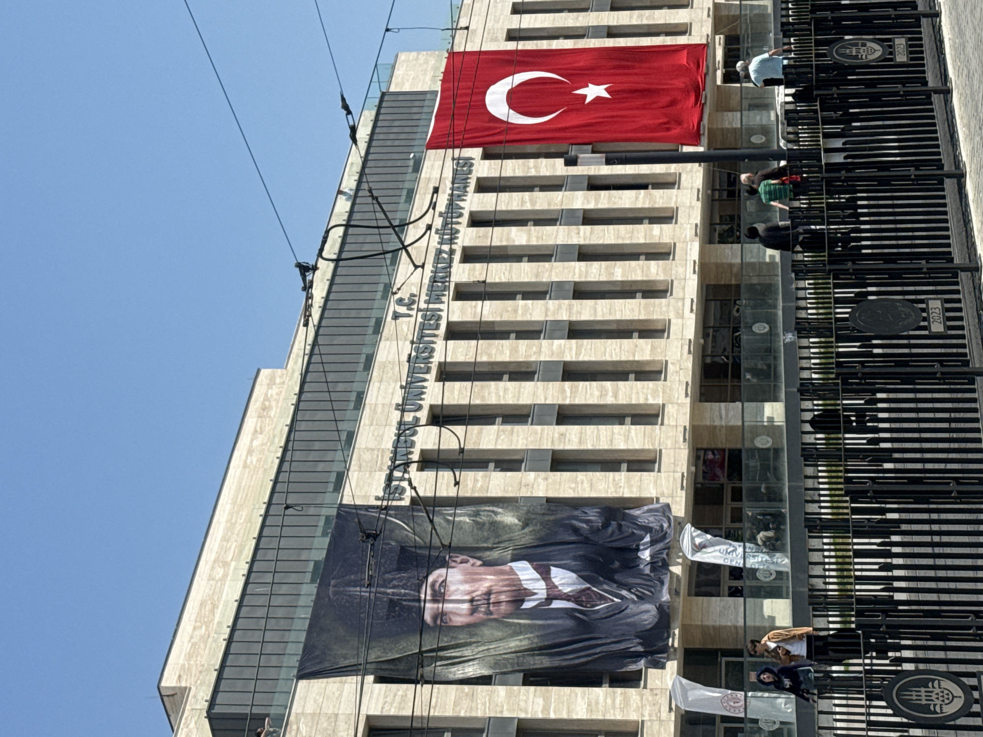 The image depicts the facade of a building in an urban setting, identified by the inscription on the building as "T.C. İSTANBUL ÜNİVERSİTESİ MERKEZ KÜTÜPHANESİ". This inscription indicates it is a central library of Istanbul University.
Hanging from the building are two prominent items: a large Turkish flag featuring the white crescent and star, and a large banner with a portrait of Mustafa Kemal Atatürk, the founder of the Republic of Turkey.
A wrought iron fence runs vertically along the right side of the building, partially obscuring the view. People are visible behind the fence, positioned on a ledge or balcony of the building.
The sky is a clear, light blue. The building itself appears to be constructed of stone or a light-colored material, with rows of windows across multiple floors. Electrical or communication wires stretch across the top portion of the building in the foreground.