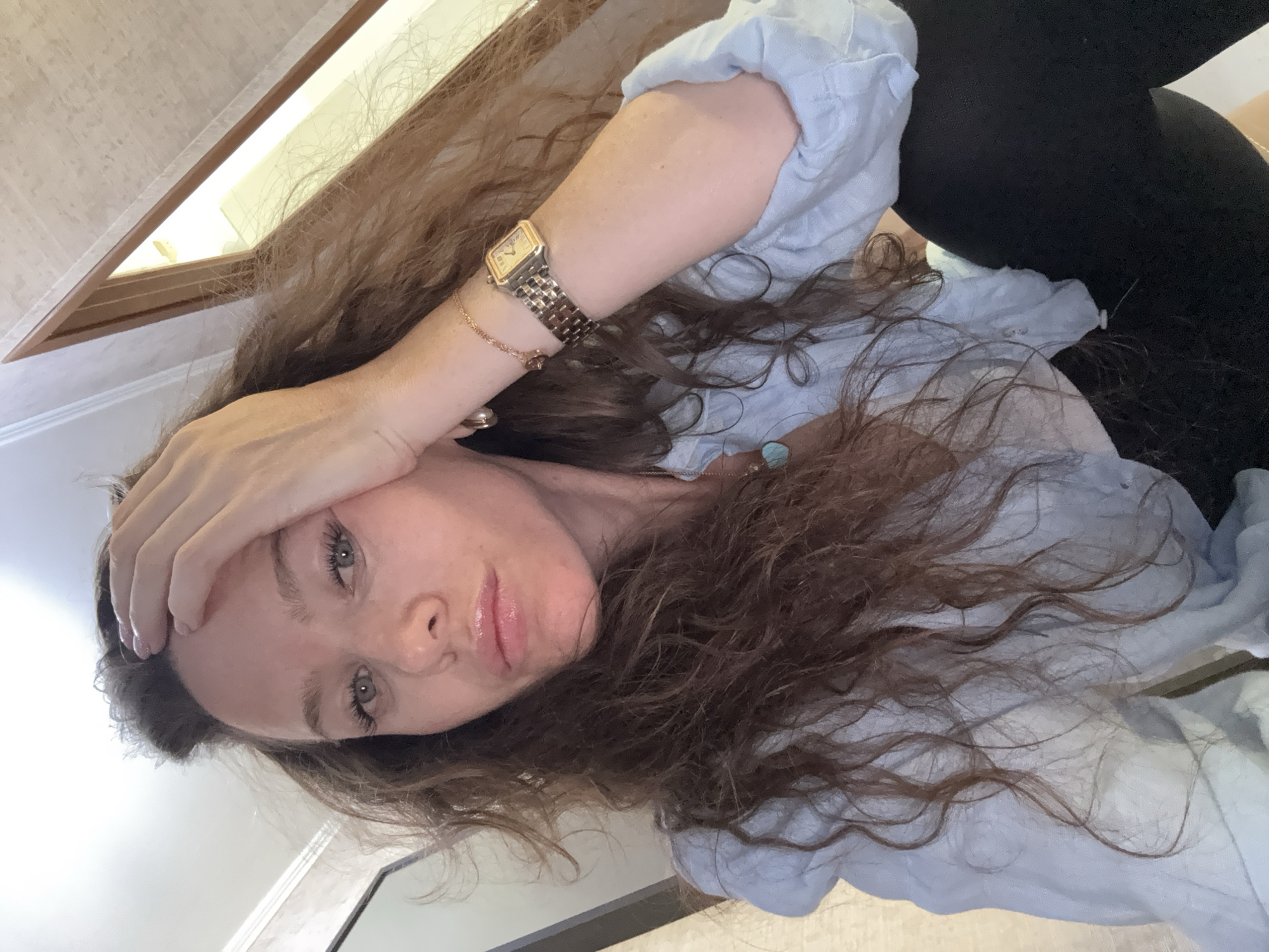 The image shows a young woman in an indoor setting, posing for what appears to be a selfie. Her head is tilted, and she looks directly at the camera with light green or hazel eyes. She has long, wavy brown hair that falls around her face and shoulders. Her right hand rests on her forehead in a casual or contemplative gesture. Her lips are glossy, and her complexion is clear.
She is wearing a light blue, possibly linen, button-up shirt with rolled-up sleeves, and part of dark clothing, possibly leggings or trousers, is visible. On her left wrist, she wears a gold-toned rectangular watch and a delicate gold bracelet. A small turquoise pendant hangs from a necklace around her neck.
The background reveals a light-colored, textured wall. In the upper left corner, a bright light source, possibly a window or a ceiling light fixture, is visible, framed by a wooden border. The lighting suggests an indoor environment, either during the day or in a well-lit room. The overall mood conveyed by her expression is relaxed or slightly pensive. There is no visible text in the image. The location context provided is Moscow, Russia.