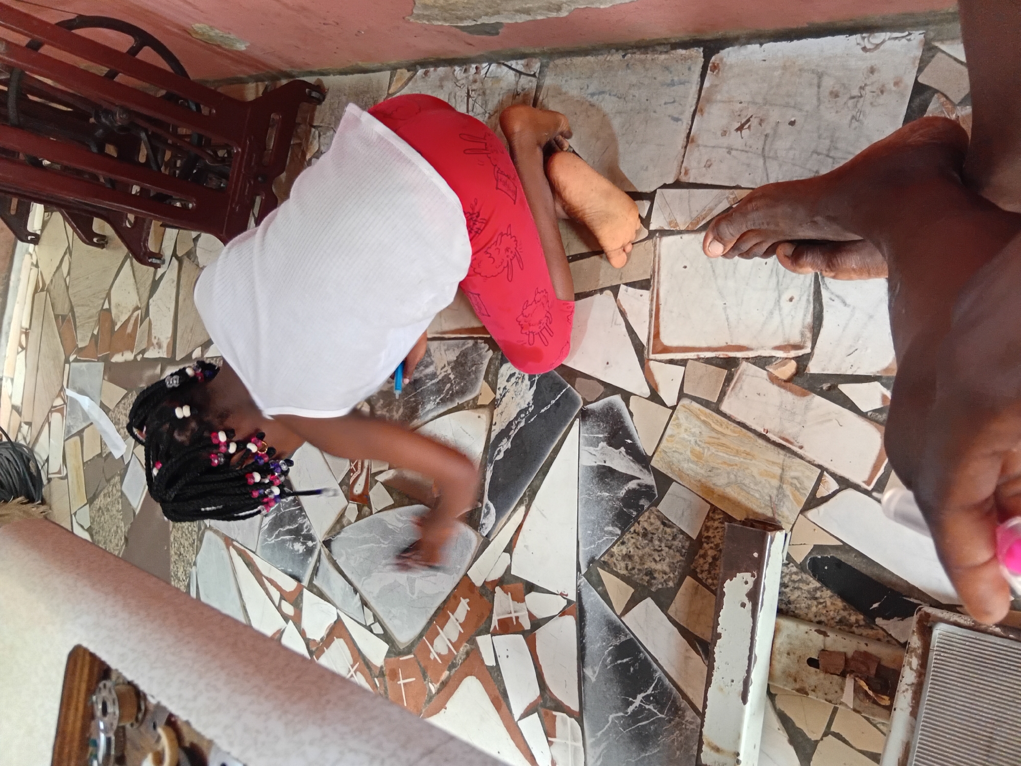 A person is kneeling on a tiled floor and wiping it with a cloth. The person is wearing a white tank top and red leggings with a cartoonish print. The person's hair is braided with colorful beads. In the background, there is a vintage sewing machine and a wall with peeling paint. The floor is made of irregularly shaped tiles in various colors and patterns. Another person's feet are visible in the foreground, suggesting the image is taken from a low angle. The scene appears to be indoors, possibly a home environment.