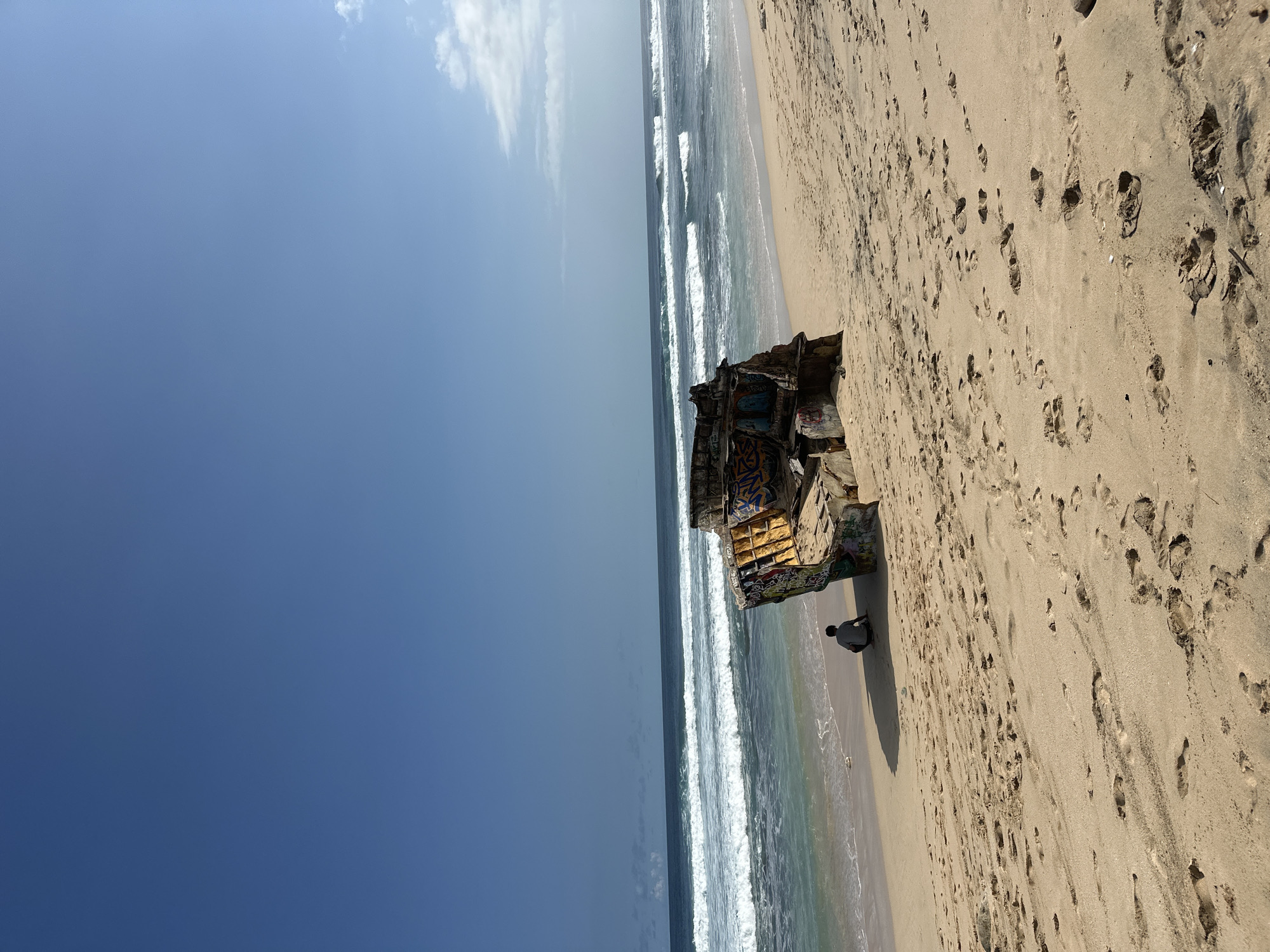 The image shows a dilapidated, graffiti-covered structure on a sandy beach in Kuta Selatan, Indonesia. The building's skeletal remains stand partially submerged in the ocean, with waves lapping at its base. A single person is sitting near the structure, seemingly contemplating the scene. The beach itself is marked with numerous footprints, suggesting prior human activity. The sky is a clear blue, contrasting with the weathered appearance of the building and the dynamic movement of the waves.