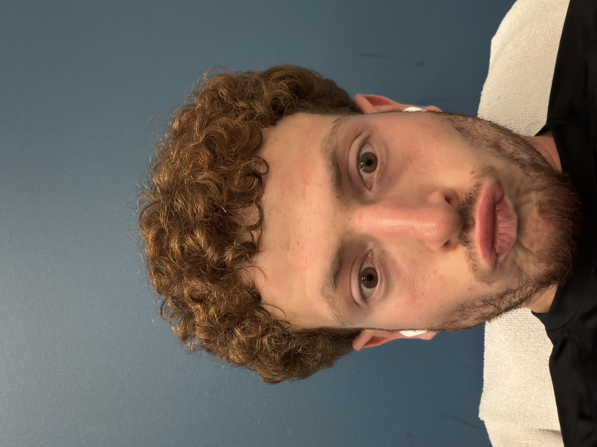 A close-up, head-on, selfie-style photograph features a person with curly, auburn hair and light brown eyes. The person is wearing white earbuds and has a light beard. They are puckering their lips, and a white towel is visible behind their shoulders. The background is a plain, teal-colored wall. The photograph was taken in Beachwood, United States of America.