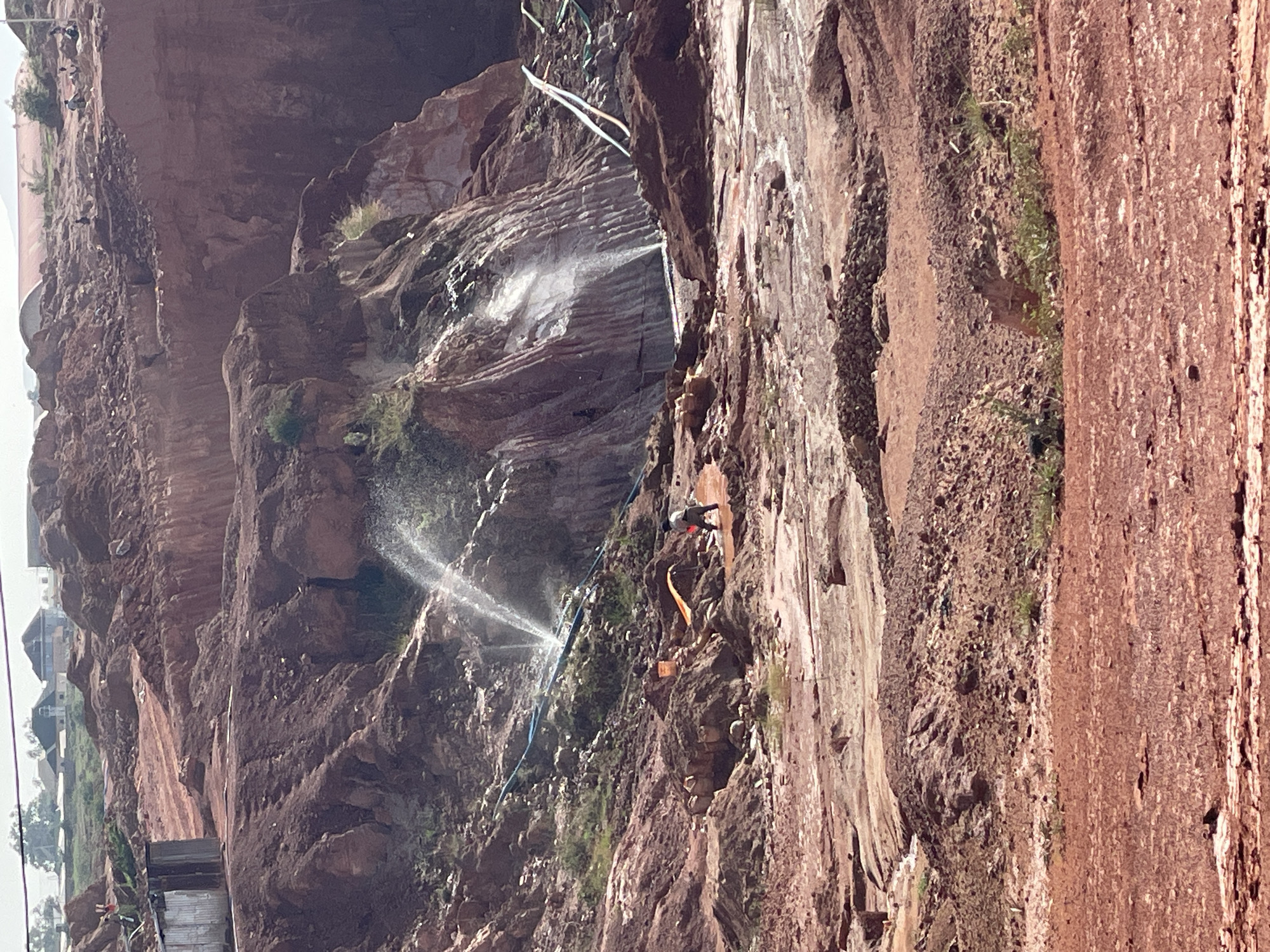A person is suspended on the side of a reddish-brown rock face, potentially a cliff or quarry, spraying water. There are multiple streams of water being directed at the rock face, and some smaller areas of vegetation are visible. Houses are visible in the distance, suggesting this might be located in an urban area of Jos, Nigeria. The environment is rocky and potentially dusty. There are also pipes and other equipment present, indicating some type of industrial or construction activity.