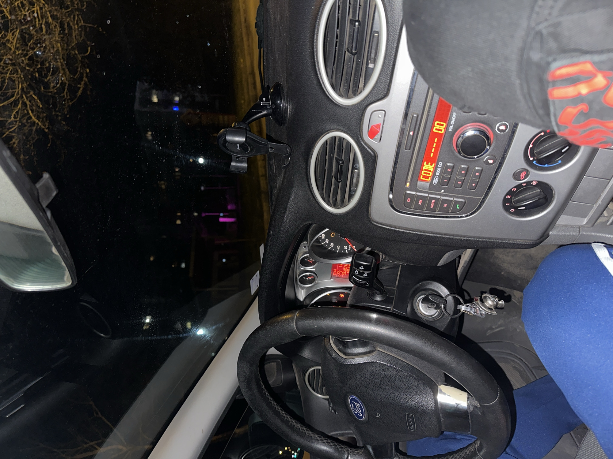 The image shows the interior of a Ford vehicle. The dashboard, steering wheel with the Ford logo, speedometer, and radio console are visible. The radio display shows the word "CODE" and the time "15:48". A smartphone holder is attached to the dashboard. The key is in the ignition. The background shows a nighttime view through the windshield, with some blurred lights and trees. The setting appears to be an urban area.
There are no features that confirm this image was taken in Varna, Bulgaria.