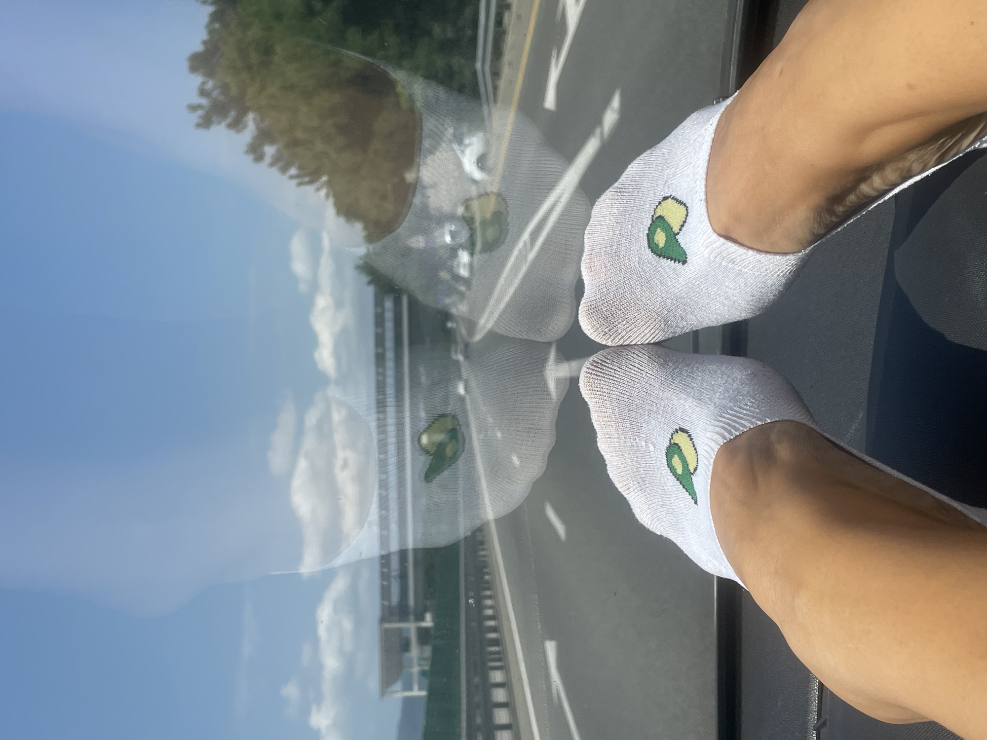 A pair of white ankle socks with a green and yellow avocado design are worn by a person whose legs and feet are visible. The socks are on the person's feet, which are resting on the dashboard of a vehicle. The person is on a highway, with a bright blue sky and scattered clouds visible above. The reflection in the windshield shows a highway with traffic, a building in the distance, and trees. The image appears to have been taken during the day.
