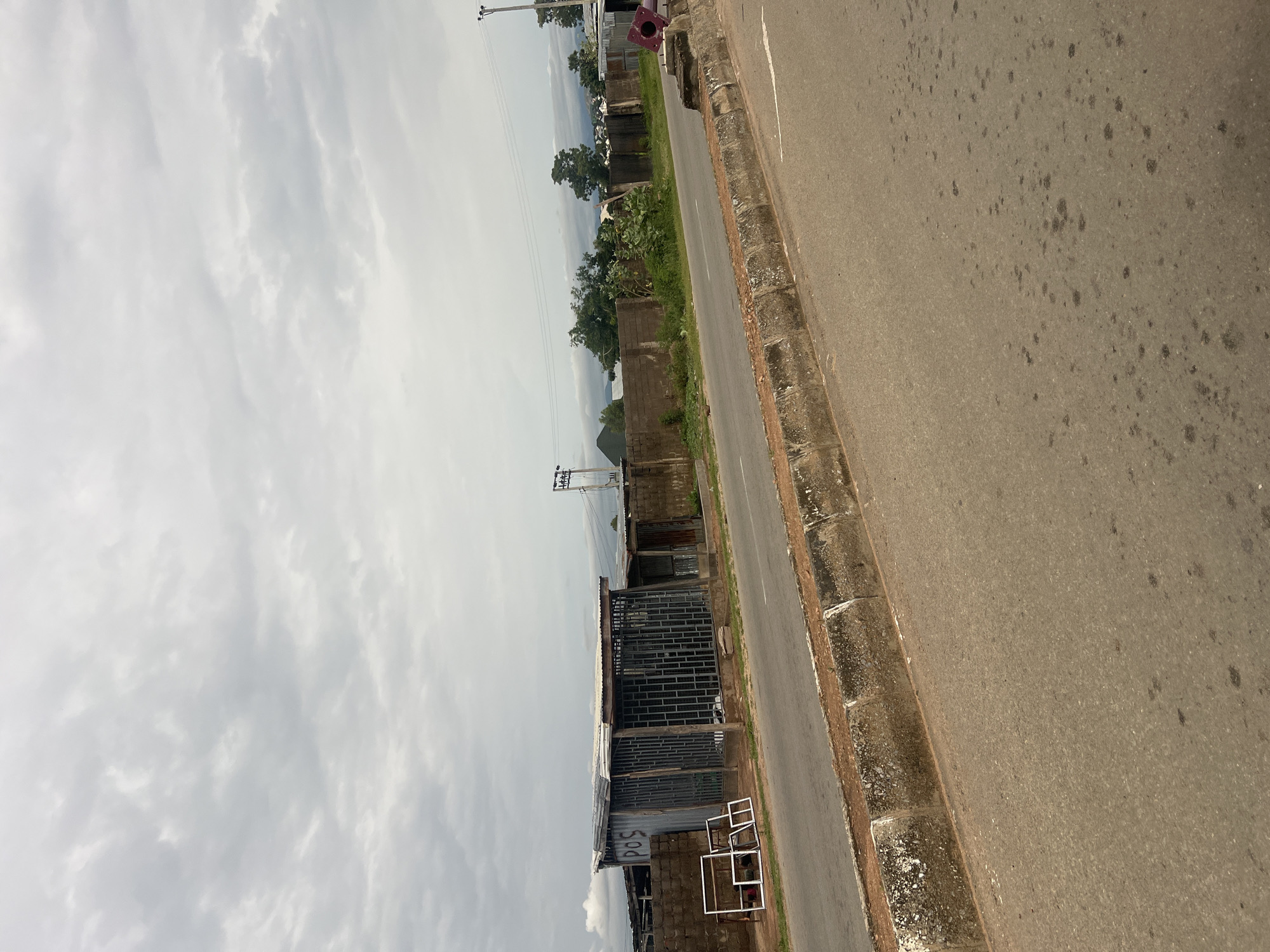 The image depicts a roadside scene in Jalingo, Nigeria. The viewpoint is from an elevated position, looking down at an angle. The foreground is dominated by a gray asphalt road with a concrete curb. The road shows signs of wear, with dark stains scattered across its surface.
To the left of the road, a narrow strip of green grass separates it from a row of buildings and structures. These structures appear to be a mix of brickwork, corrugated metal roofing, and metal grates, suggesting a commercial or residential area. One building has a sign with faded red lettering that might read "POS." There are several white square frames arranged on the ground in front of one of the buildings. Further down the road, trees and vegetation are visible, interspersed with more buildings and a utility pole with wires. The background shows a hazy sky with visible clouds and the suggestion of distant hills or elevated terrain.
The overall impression is one of a typical day in a developing area. The lighting suggests it is daytime, possibly overcast or late afternoon, given the muted light and the lack of strong shadows. The weather appears to be cloudy. There are no discernible people actively engaged in an activity, but the presence of buildings and the road indicates a populated area. The scene is static, with no apparent event unfolding.