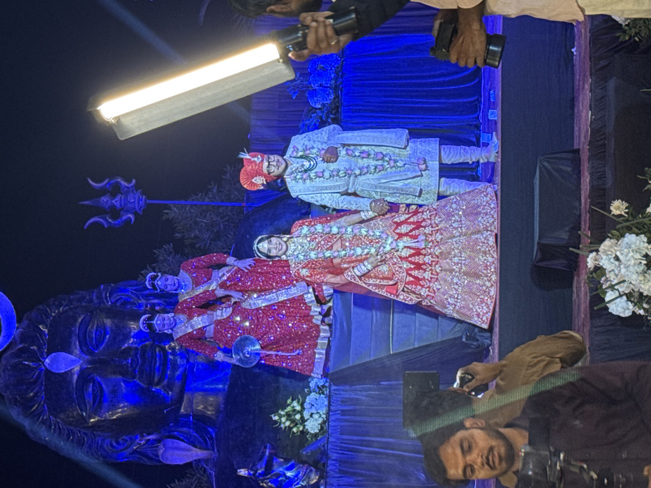 A wedding scene unfolds with a bride and groom in traditional attire, flanked by statues of Hindu deities. Photographers capture the moment, one with a light bar. The groom wears a light-colored suit and a turban, while the bride is adorned in a red and gold lehenga. Garlands decorate both. A large sculpture of Shiva's head, bathed in blue light, dominates the foreground. A trident symbol stands tall behind the couple. The backdrop is draped in blue fabric, embellished with white floral arrangements.