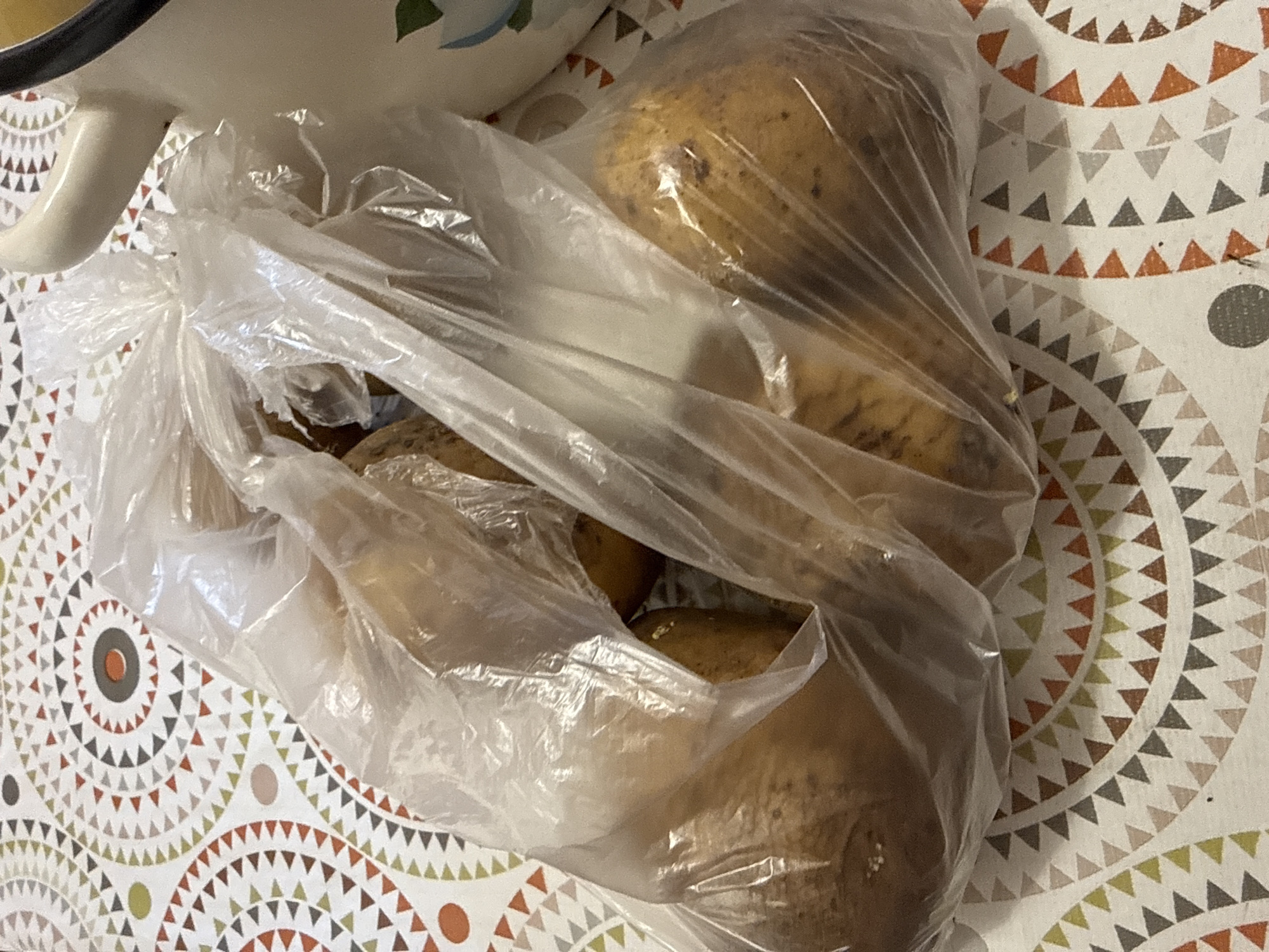 The image shows a plastic bag filled with potatoes resting on a patterned tablecloth. The tablecloth has a repeating geometric design featuring triangles and circles in shades of white, cream, orange, brown, green, and gray. Part of a white ceramic bowl or pitcher with a black rim is visible in the upper left corner of the image, suggesting it might be used to hold or serve the potatoes. The potatoes themselves are light brown and appear to be raw, with some dirt or imperfections visible on their skins. The lighting suggests it is daytime, and there are no specific cues to indicate the weather or a particular emotion. The scene appears to be indoors, likely in a kitchen or dining area. There is no visible text in the image. The location context of Morozovka, Russia is provided, but there are no visual elements within the image that specifically confirm this location.