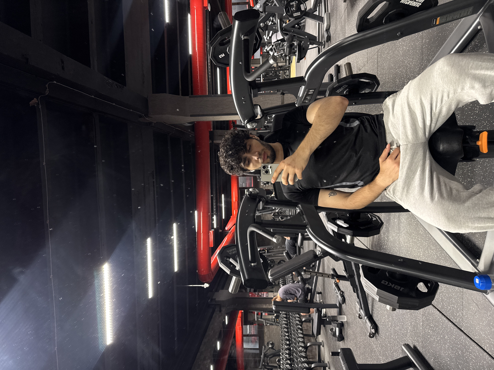 A young man with curly dark hair is sitting on a weight machine in a gym, taking a selfie with his phone. He is wearing a black t-shirt and grey sweatpants. The gym appears to be modern and well-equipped, with rows of dumbbells and other weight machines visible in the background. Red pipes run along the ceiling, and fluorescent lights illuminate the space. The floor is a dark rubber matting. The scene is likely in Yerevan, Armenia, given the context of previous requests.