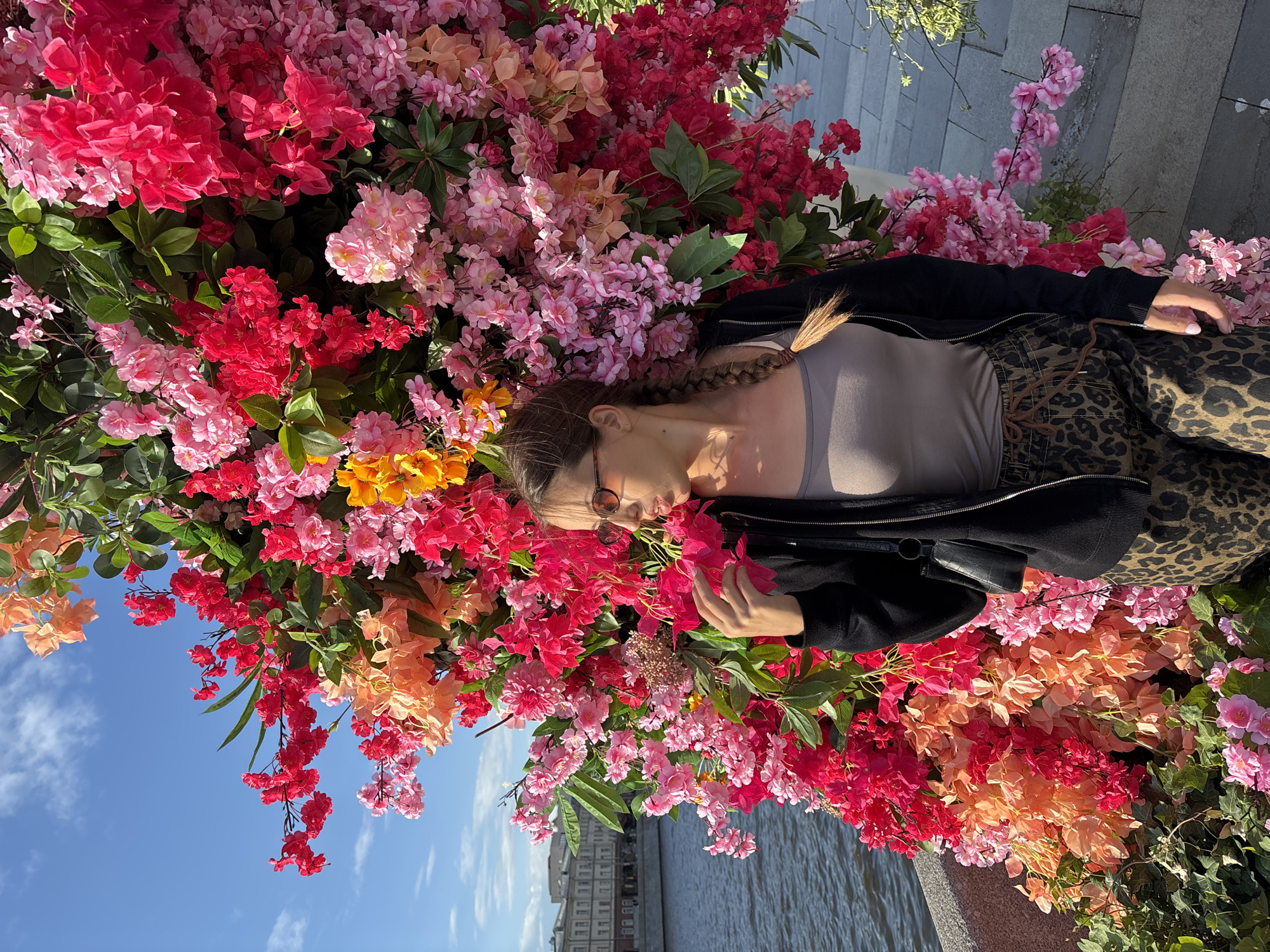 A woman is lying amongst a profusion of artificial flowers in a public space in Moscow, Russia.
The woman has fair skin, wears glasses and has long brown hair styled in a braid. She wears a light-colored top, a black jacket, and patterned pants. She is lying amongst a large arrangement of artificial flowers of varying shades of red, pink, and orange. Some green foliage is visible among the flowers.
In the background, a river can be seen, and a row of buildings is visible along the riverbank. The sky is blue with a few wispy clouds. The scene appears to be outdoors during daylight hours.