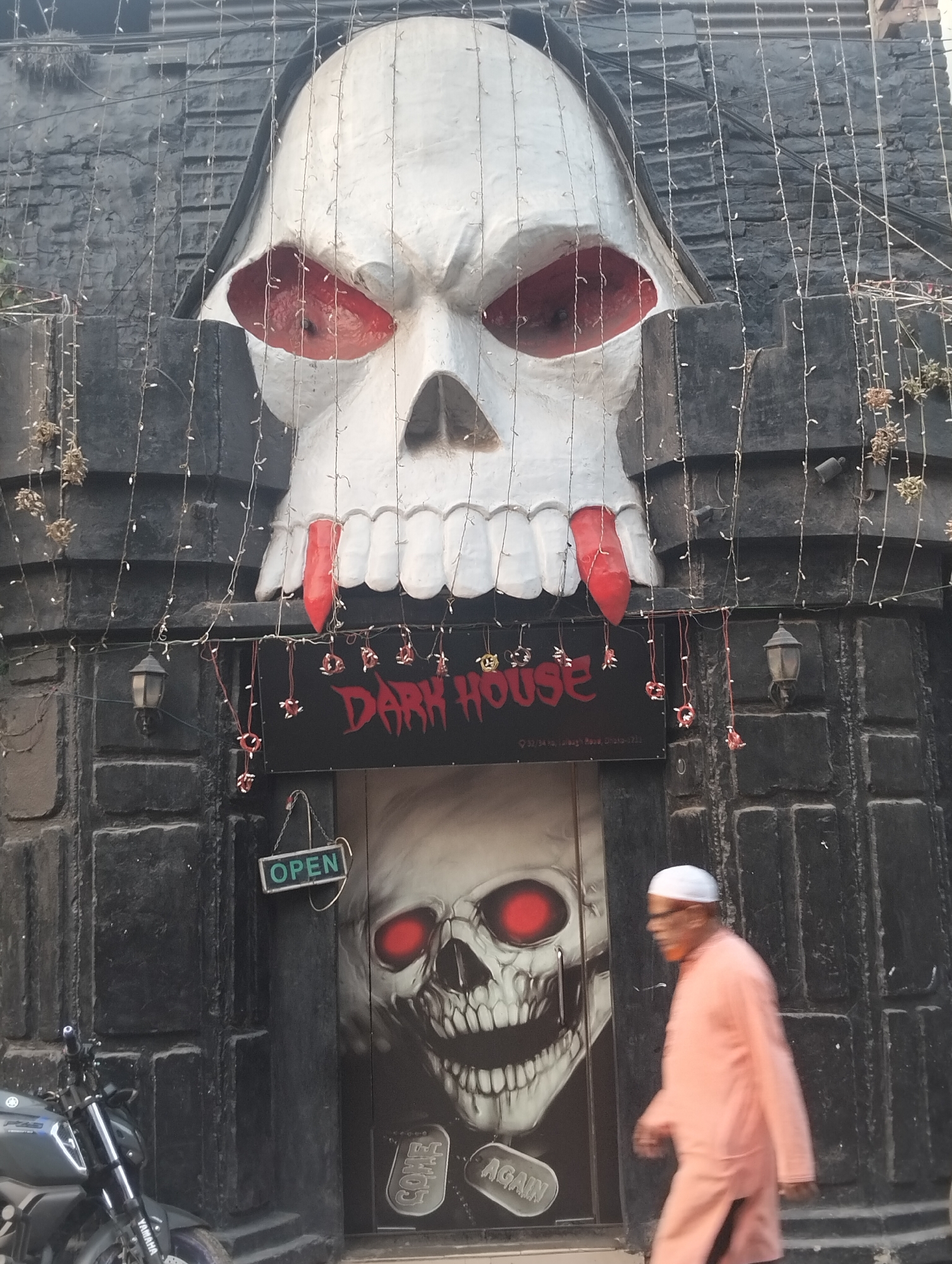 The image depicts the entrance of "DARK HOUSE", a horror-themed establishment located at 33/34 Khajeh Reta Dhaka-1211, Bangladesh. Above the entrance is a large, white skull with red eye sockets and what appear to be red blood drips from the teeth. The building's facade is dark gray, resembling a castle-like structure, and decorated with hanging lights. Below the sign, the door also features a skull motif, with red glowing eyes, and text "COME AGAIN". An elderly man wearing a white cap and peach-colored clothing is walking past the entrance. A motorcycle is parked in front of the building to the left. An "OPEN" sign is hanging in the doorway.