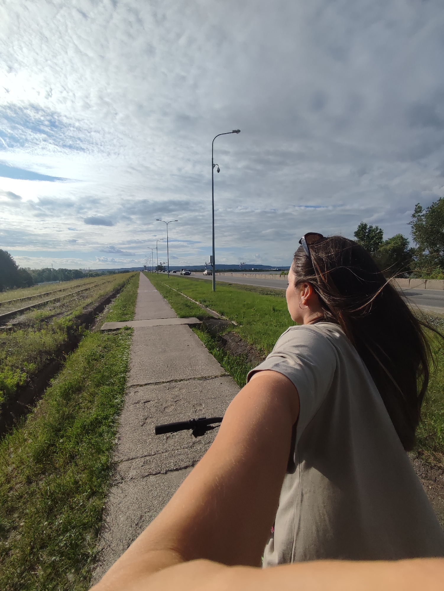 In this selfie-style video, a woman is captured from the chest up, riding a bicycle on a paved path next to a set of disused railway tracks. She is wearing a light grey t-shirt and sunglasses are perched on top of her head. Her long, dark hair is blowing in the wind. The path appears to be a pedestrian or bike lane alongside a road with traffic visible in the distance. The setting is outdoors with green grass on either side of the path and tracks. The sky is partly cloudy with a mix of bright and overcast conditions, suggesting it's daytime. The mood appears relaxed and active, as the woman is enjoying a bike ride outdoors.