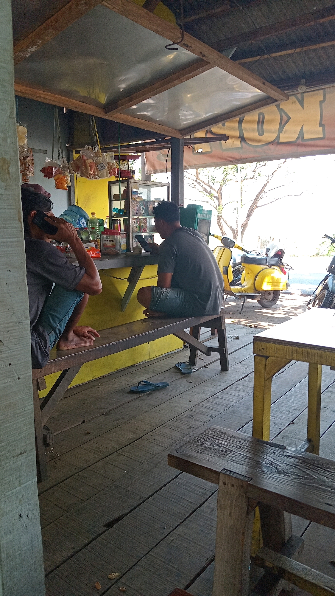Two men are seated on a wooden bench in a covered outdoor area with a wooden floor. The man on the left, facing away from the camera, is wearing a grey t-shirt and blue jeans and is holding a mobile phone to his ear. The man on the right, also facing away from the camera, is wearing a grey t-shirt and blue shorts and is looking at a mobile phone. A yellow Vespa scooter is parked behind them, and other motorcycles are visible in the background. The scene appears to be a small roadside stall or warung in Ambeng Ambeng Watangrejo, Indonesia, indicated by the presence of various packaged goods and food items displayed on shelves behind the bench. The overall environment is rustic and informal, with a bright yellow wall behind the counter.