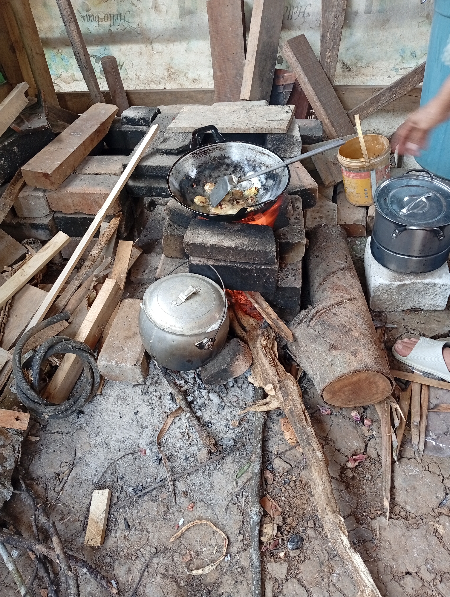 The image depicts an outdoor cooking scene on a dirt floor. In the center, a rustic stove constructed from bricks is in operation, with a wok over a fire containing food being stirred by a spatula. To the left of the stove, a metal pot is placed on the ground. To the right, a large log is situated near a person's foot wearing a white sandal. A yellow bucket is visible behind the wok. Various pieces of wood and debris are scattered around the area. The background shows a wall with some writing. The scene appears to be in Cisitu, Indonesia, suggesting a rural or traditional setting.