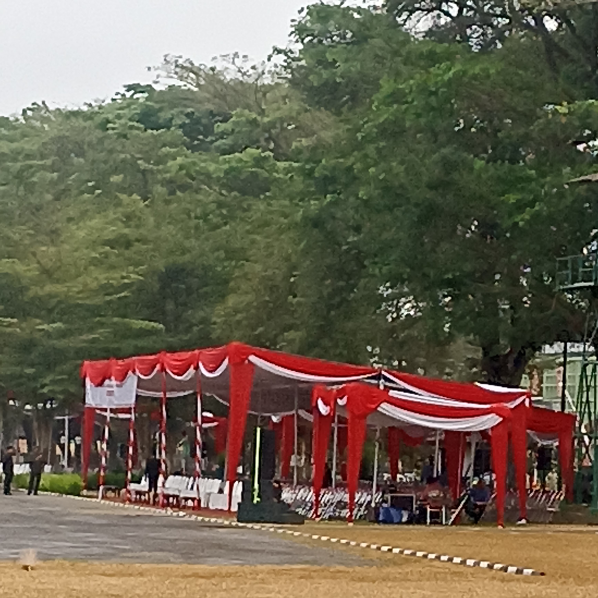 This is a medium-shot, eye-level photograph taken outdoors on a hazy day. In the foreground, a dry, yellow-brown grassy field is visible, with a paved area beyond it. A black and white striped border runs horizontally across the paved area.
Dominating the mid-ground is a large, open-sided tent with a red and white canopy. Red drapes hang decoratively from the edges of the tent. Underneath the tent, several white chairs are arranged in rows. Tables are also present, some with blue items on them. People are visible inside and around the tent; some are standing, and others are seated.
In the background, dense green foliage from numerous trees forms a thick wall of greenery. Partially visible behind the trees is a tall, green metal structure, possibly a stage or viewing platform. Several people in dark clothing are standing on the paved area to the left of the tent, facing towards it. One person is wearing a black jacket and dark pants, while another is in a dark uniform.
The overall impression is of an outdoor event or gathering, possibly a ceremony or festival, taking place in a location with abundant trees. The hazy sky suggests overcast or humid weather. Given the context of the photo being from Magelang, Indonesia, this scene could depict a local community event, a school function, or a military ceremony, which are common occurrences in the region.