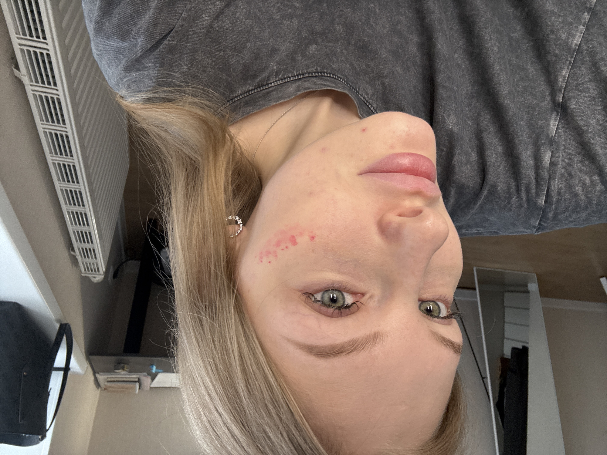 A close-up, rotated portrait shot of a fair-skinned young woman is presented. The woman has light blonde hair, green eyes, and appears to have some skin imperfections, including small red blemishes on her forehead and cheek. She is wearing a gray T-shirt and a delicate necklace with a small pendant. The background reveals a partial view of a room with a radiator, a dark-colored handbag, a small table with objects, and a glimpse of a mirror. The location is not discernible.