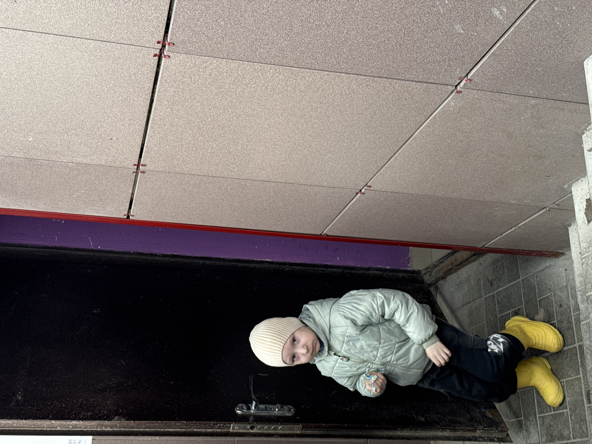 A full-body, eye-level shot captures a young child standing in what appears to be an indoor hallway or entrance area. The child is dressed warmly in a light green quilted jacket, dark pants, and a light-colored ribbed beanie, complemented by bright yellow, possibly rubber, boots. They are looking directly at the camera with a neutral to slightly curious expression, holding a small, colorful, crinkled object, possibly a sweet wrapper or toy, in their left hand.
The child stands on a floor made of gray, rectangular tiles arranged in a grid pattern. To their left is a tall, dark, possibly wooden or metal, surface, resembling a door or a large utility cabinet, featuring a metallic door handle or latch mechanism positioned centrally. Above this dark surface, a horizontal band of reddish-purple painted wall is visible, with a thin red stripe running along its lower edge. The ceiling above is composed of light gray, speckled acoustic panels, also arranged in a grid. Small, red plastic tile spacers are visible at the intersections of the panel seams, suggesting a specific installation method for the drop ceiling. The overall lighting is bright and even, indicating an interior space during the day, possibly illuminated by artificial light. There is no visible text in the image. The setting is in Perm, Russia.