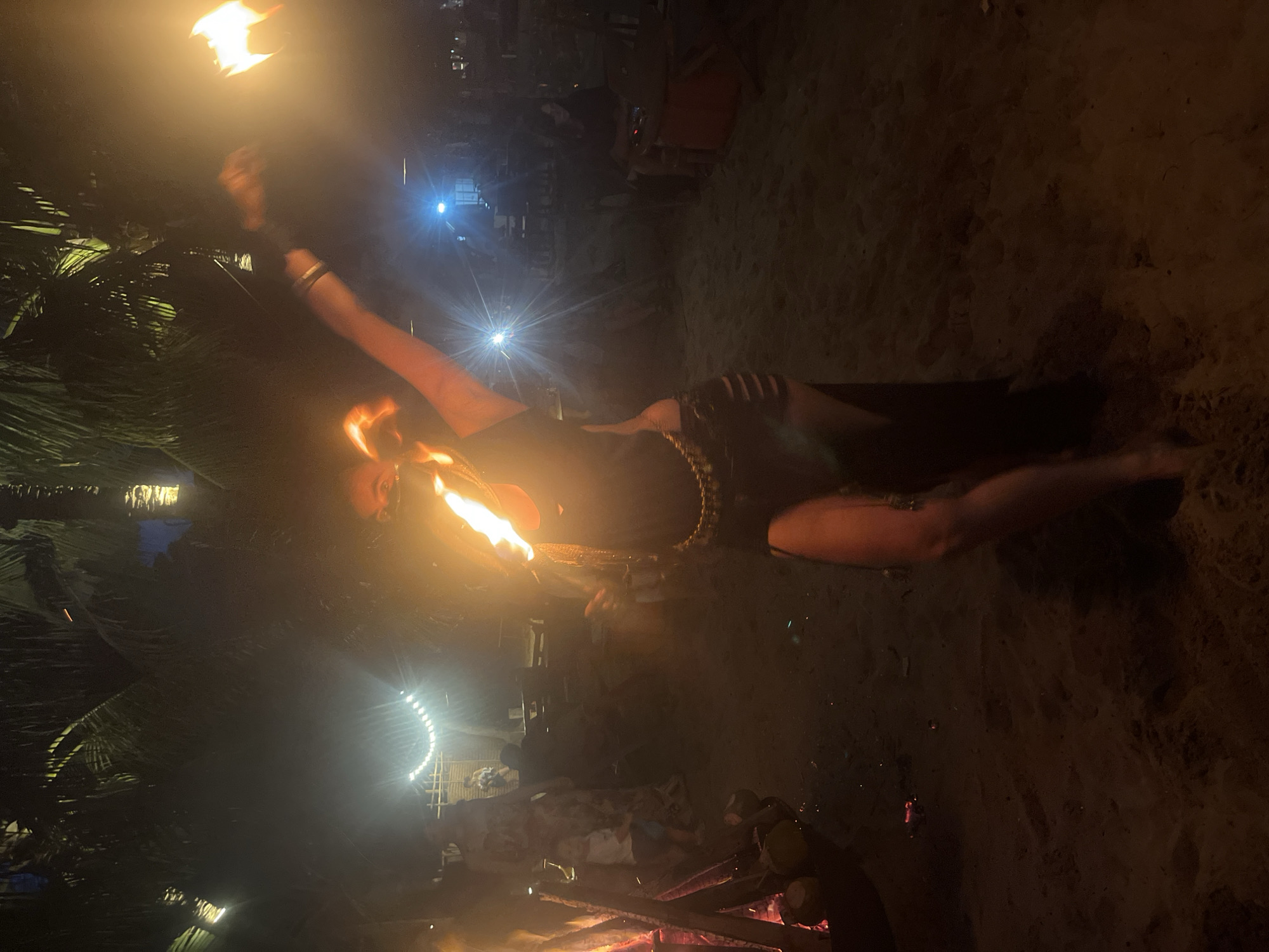A woman performs a fire breathing act on a sandy beach at night. She is wearing a dark outfit with cutouts and jewelry around her waist. She holds a flaming object in her right hand and appears to be breathing fire from her mouth, as evidenced by the flames near her face. In the background, there is a bonfire to the left, palm trees, and string lights. Several people are visible in the background, some seated and others standing. The overall scene is dimly lit by the fire and ambient lighting. This event is likely taking place at a beachside venue in Đà Nẵng, Vietnam, known for its vibrant nightlife and beachfront activities.