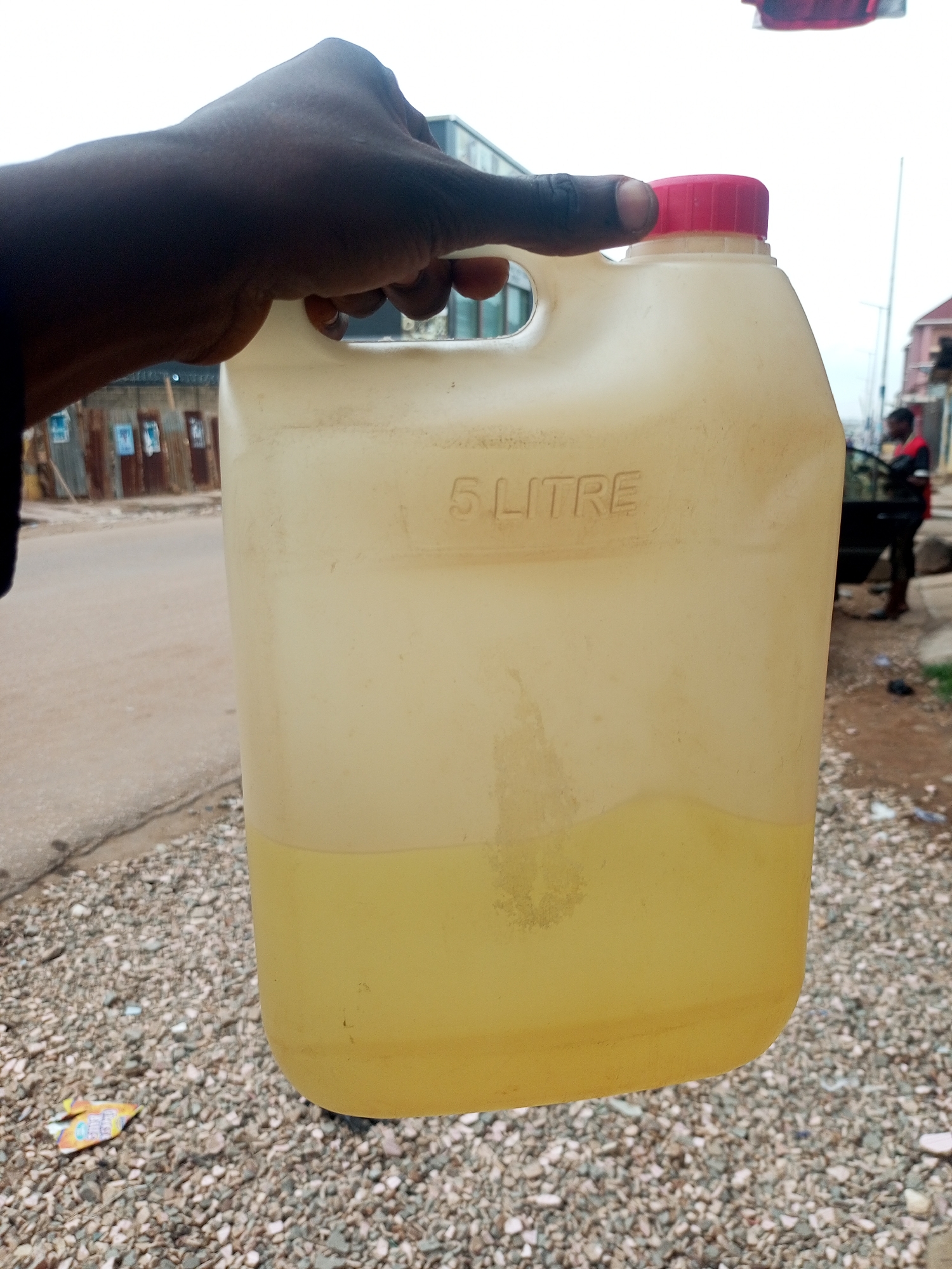A dark-skinned hand holds a partially filled, translucent yellow 5-liter plastic container with a red lid. The container appears to hold a yellowish liquid. The background shows a gravel-covered ground, a road, and buildings. A man stands next to a dark vehicle. This location is within Maiburiji, Nigeria.