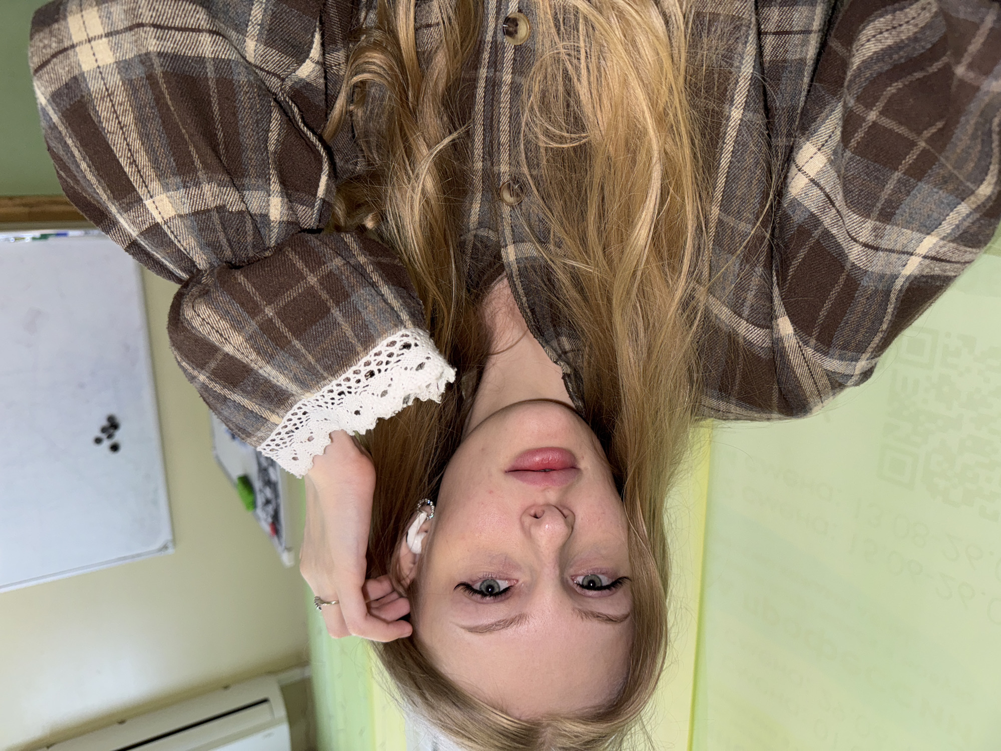 The media file features a medium shot of a young woman with blonde hair, wearing a brown and beige plaid shirt with white lace detailing on the cuffs. She is wearing a silver ring on her left hand, which is resting near her ear. She also has white earbuds in her ears. Her face is turned towards the camera, and she is looking directly at it with a neutral expression. Her makeup includes eyeliner and mascara, and her lips are slightly parted.
The background is blurred, but suggests an indoor setting. To the left, a whiteboard with some dark specks is partially visible. To the right, a pale green wall with some printed text, including QR codes, is visible. The lighting appears to be artificial and even. The orientation of the image is upside down.
The text visible on the right side appears to be in Russian and includes characters like "е" and "и", along with numbers and symbols. It is difficult to decipher fully due to the blurriness and orientation.
The overall impression is of a casual self-portrait or selfie taken indoors, likely in Russia, given the location context.